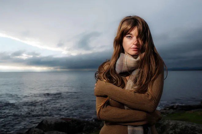A woman with long brown hair wearing a brown sweater and a beige scarf stands by a body of water with a cloudy sky in the background.