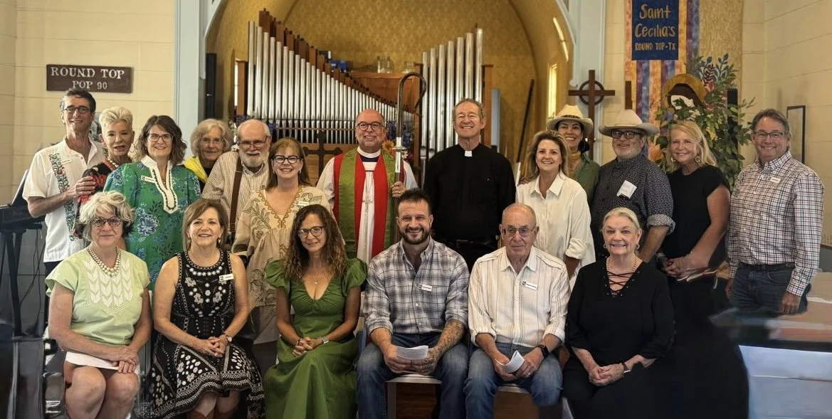 Group of people posing for a photo inside a church, with a priest in red robes holding a staff, and a black-robed clergyman. The background includes choir pipes and religious banners, one of which reads 'Saint Cecilia's Round Top-TX'.