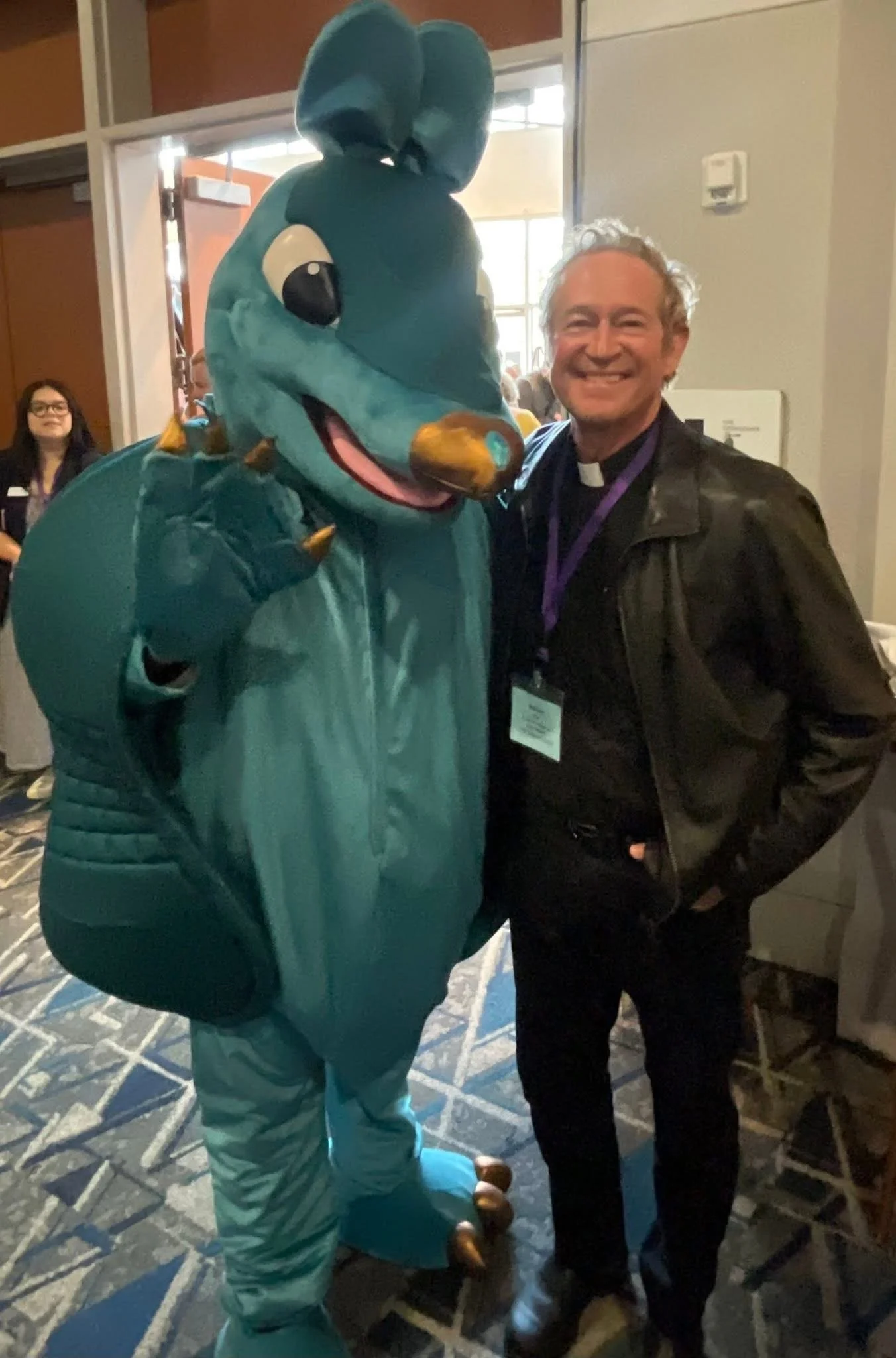 A person in a dinosaur costume posing next to a smiling man in a black jacket and clergy collar, in an indoor setting with other people in the background.
