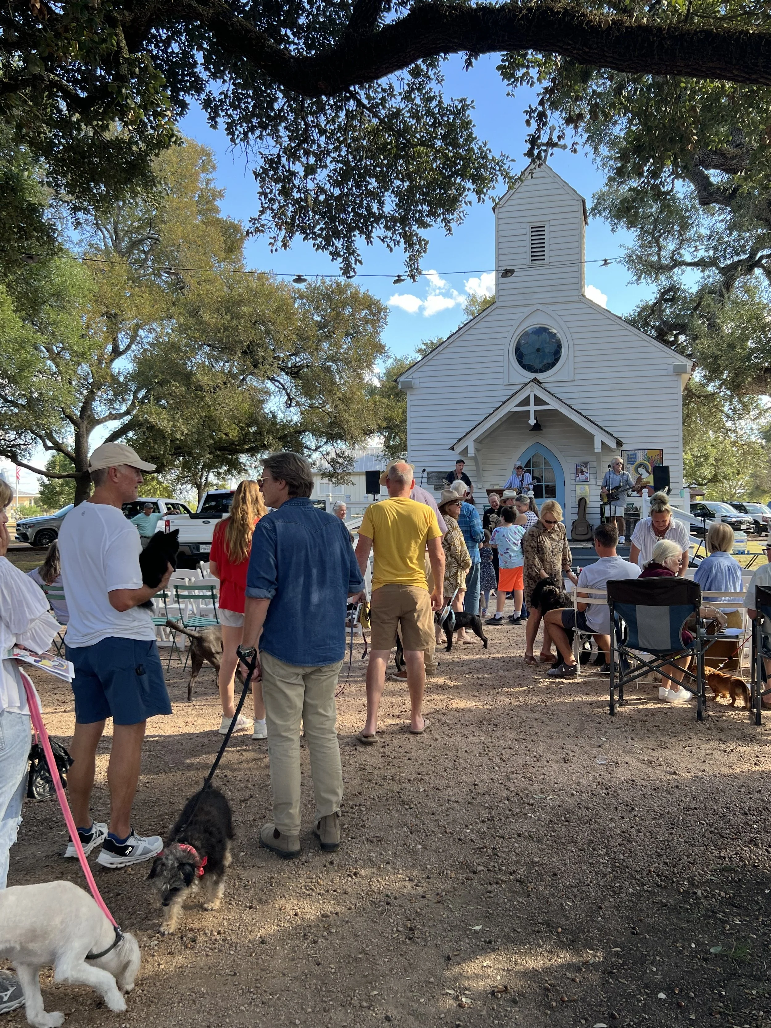 People gathered outside a small white church with a band playing on the front steps, some attendees with dogs, under large trees on a sunny day.