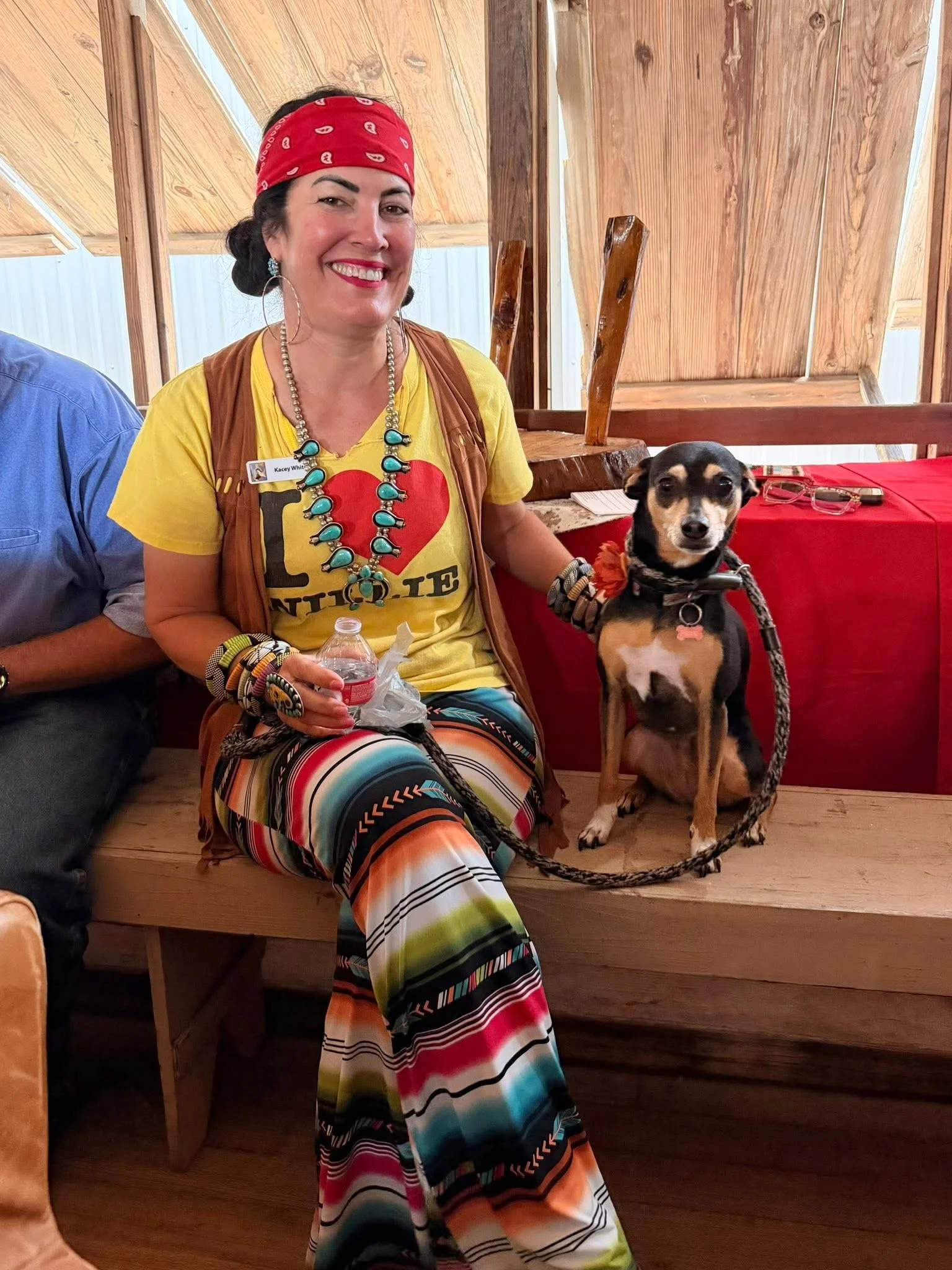 A woman with dark hair wearing a red bandana, yellow t-shirt with a red heart and the word 'LOVE' partially visible, colorful striped pants, a turquoise necklace, and large hoop earrings. She is sitting indoors on a wooden bench, smiling at the camer