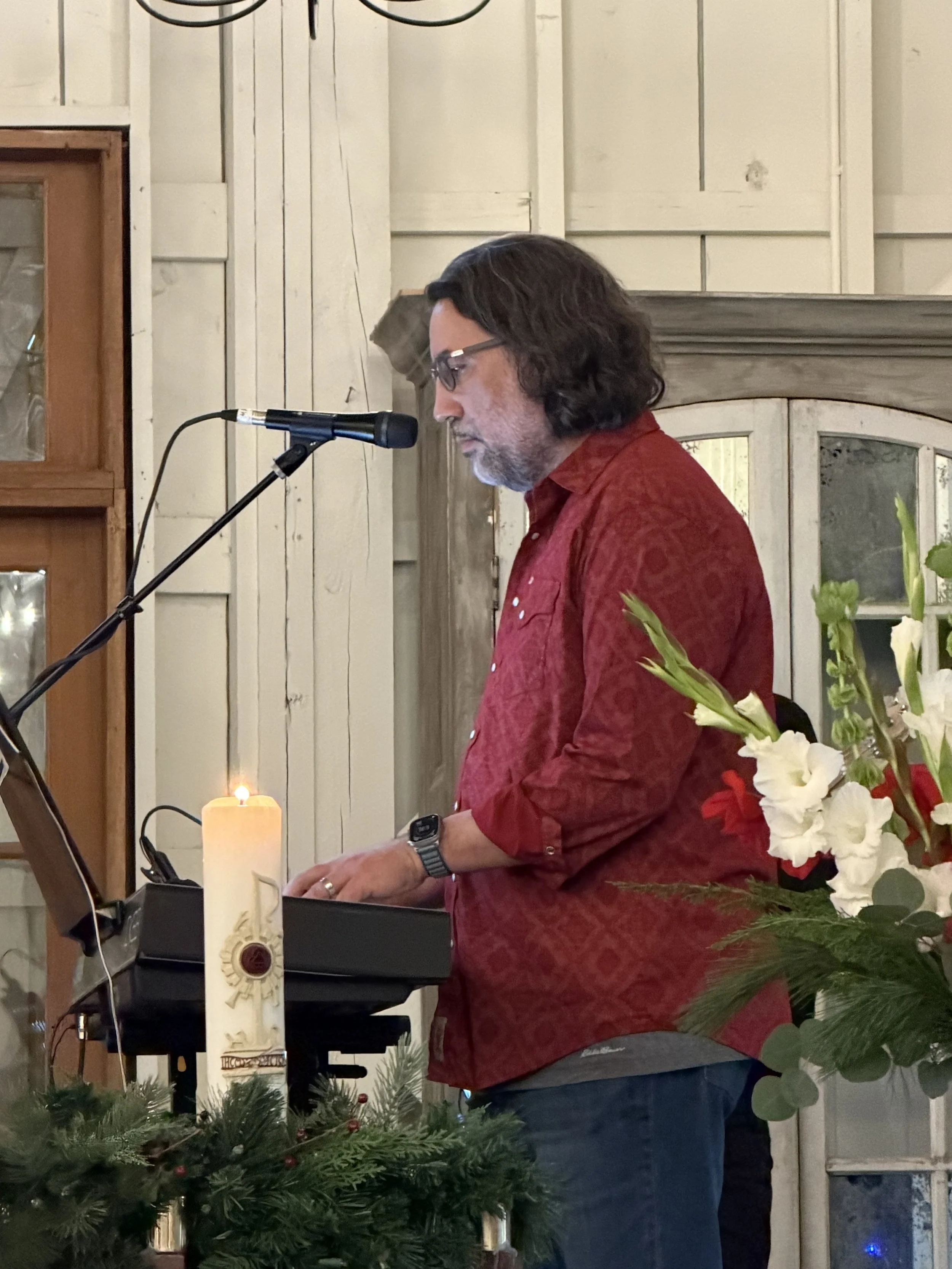 A man with dark hair and glasses, wearing a red patterned shirt, playing a keyboard at a decorated indoor event with white flowers and a lit candle, in front of a wooden wall.