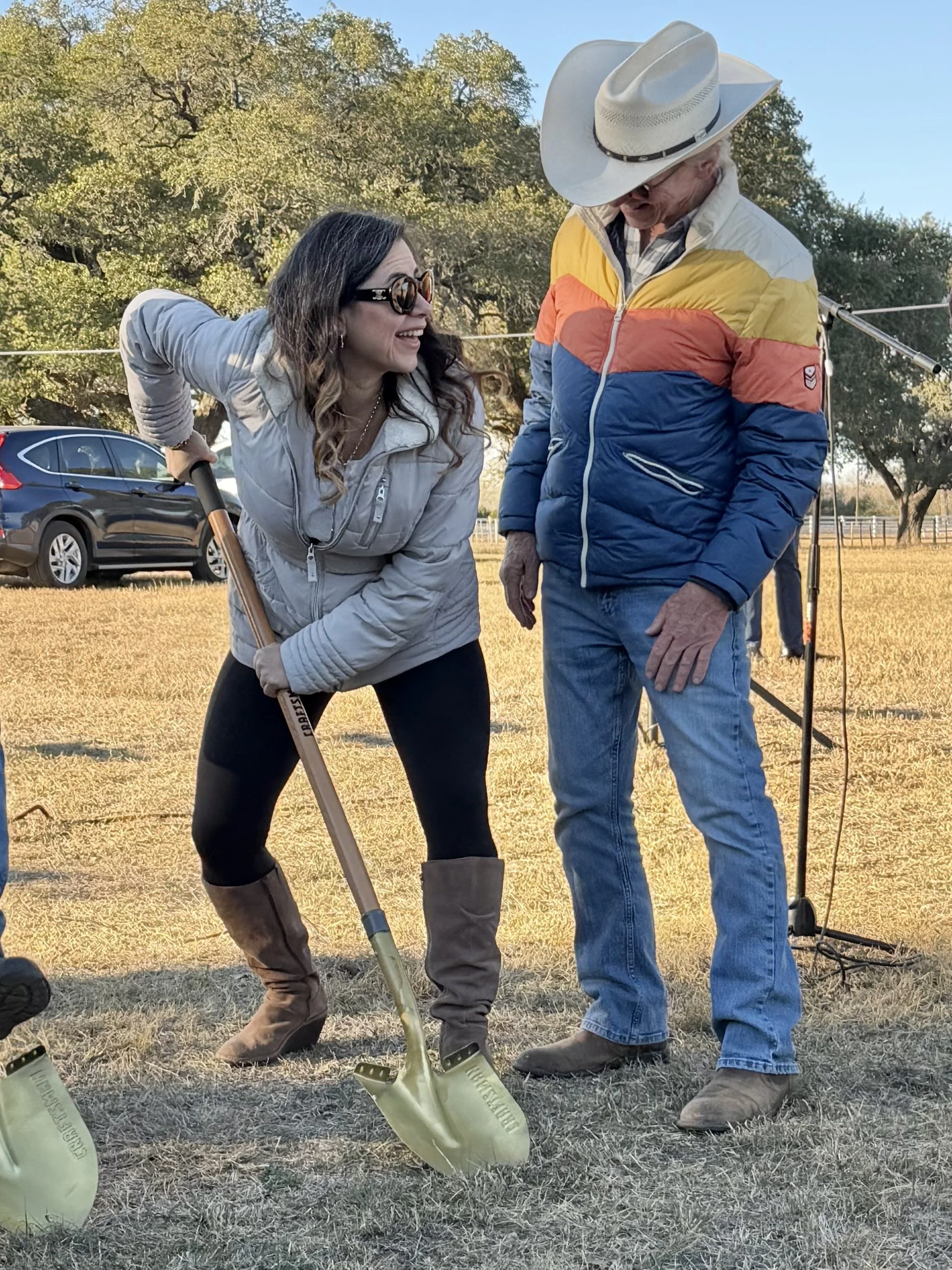 A woman with curly hair and sunglasses is smiling and leaning towards an older man wearing a cowboy hat and colorful jacket. They are outdoors, standing on dry grass, with a parked car and trees in the background. The woman appears to be holding a sh