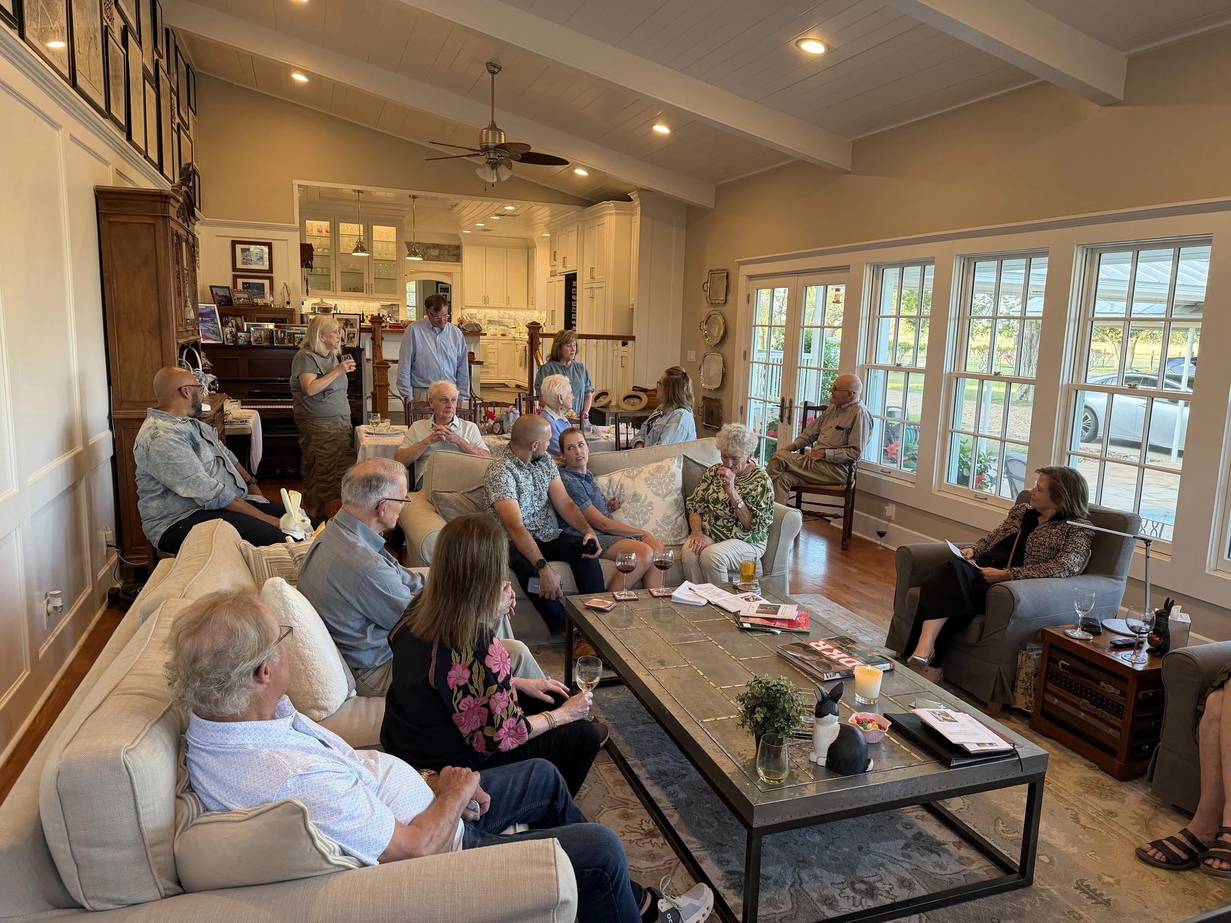 A group of people gathered in a spacious living room with large windows, hardwood floors, and a vaulted ceiling. The scene appears to be a social or family event, with some people sitting on sofas and chairs, others standing, and engaging in conversa