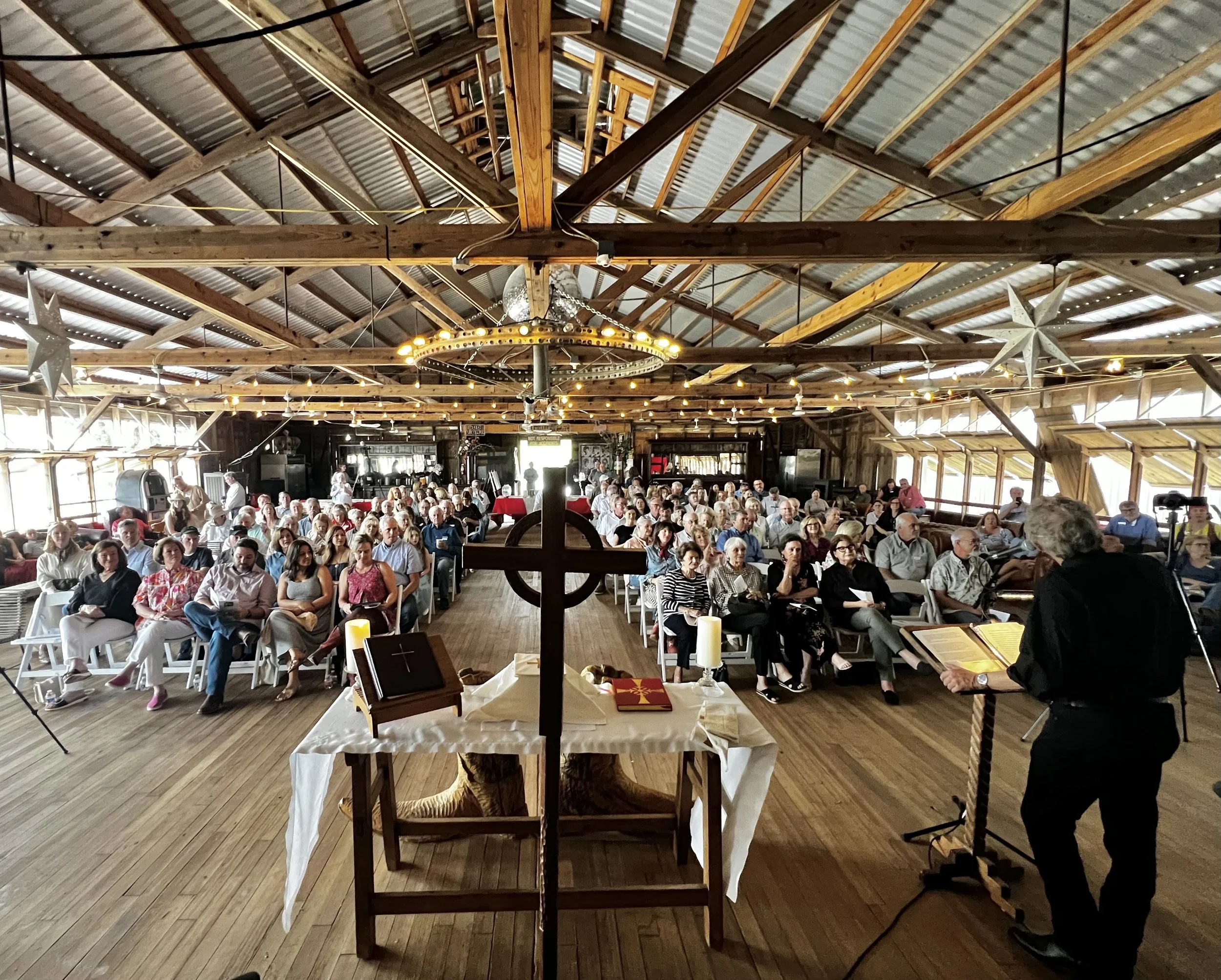Interior of a barn-style venue with a large gathering of people seated facing the stage, decorated with stars and string lights, with a cross and religious items on a table in the foreground.