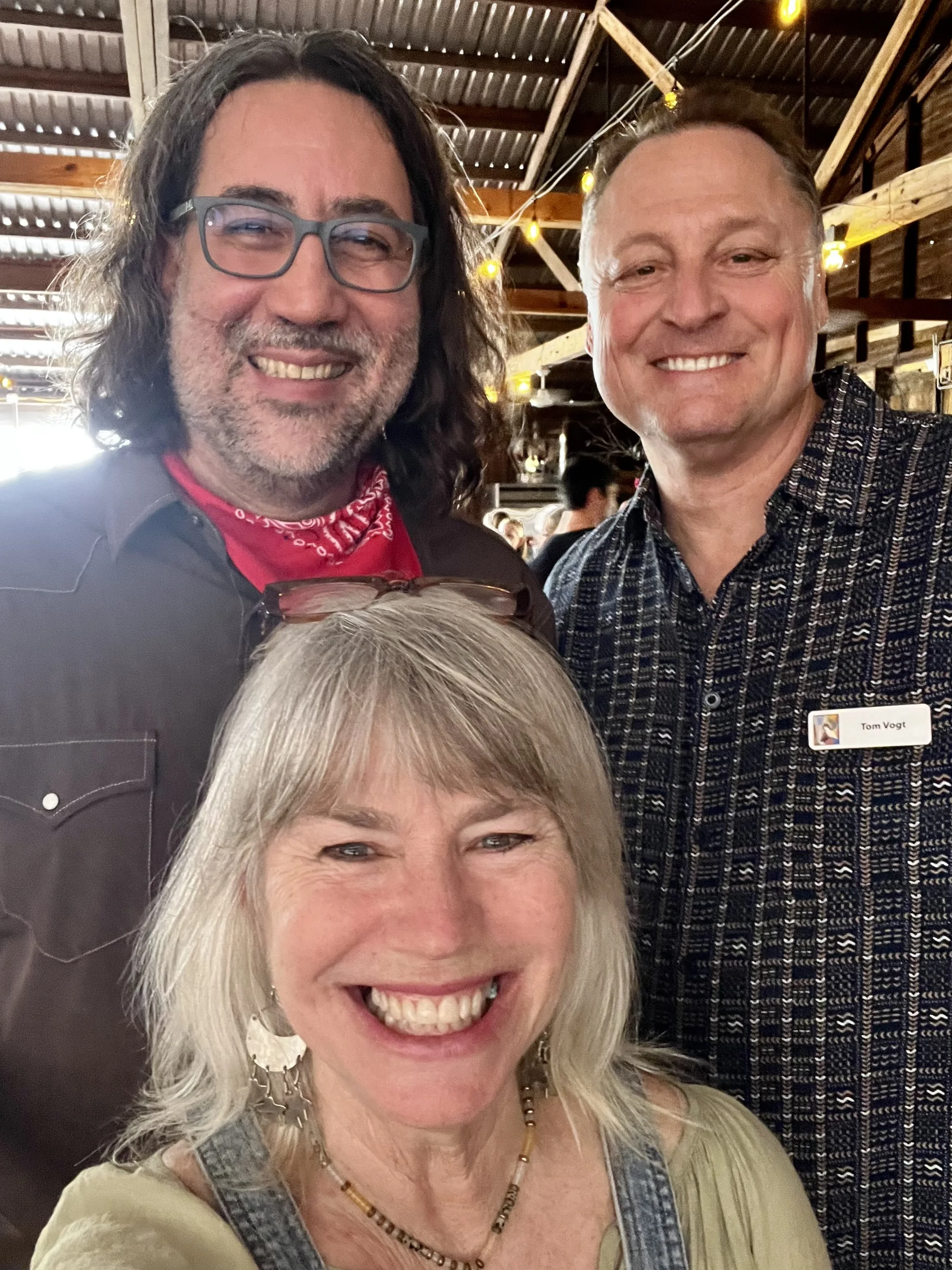 A group of three people smiling inside a rustic venue with wooden beams and string lights.