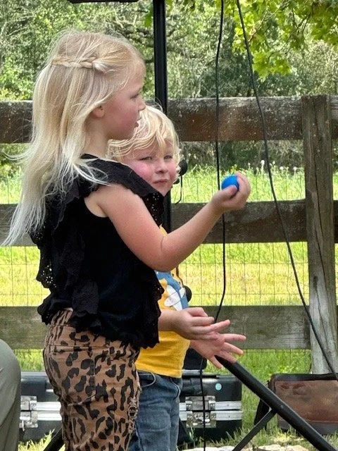 Two young children, a girl with blonde hair and a boy with light hair, playing outdoors near a wooden fence.