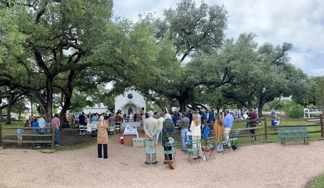 A gathering of people attending a wedding ceremony outside under large trees, with a small white chapel in the background.