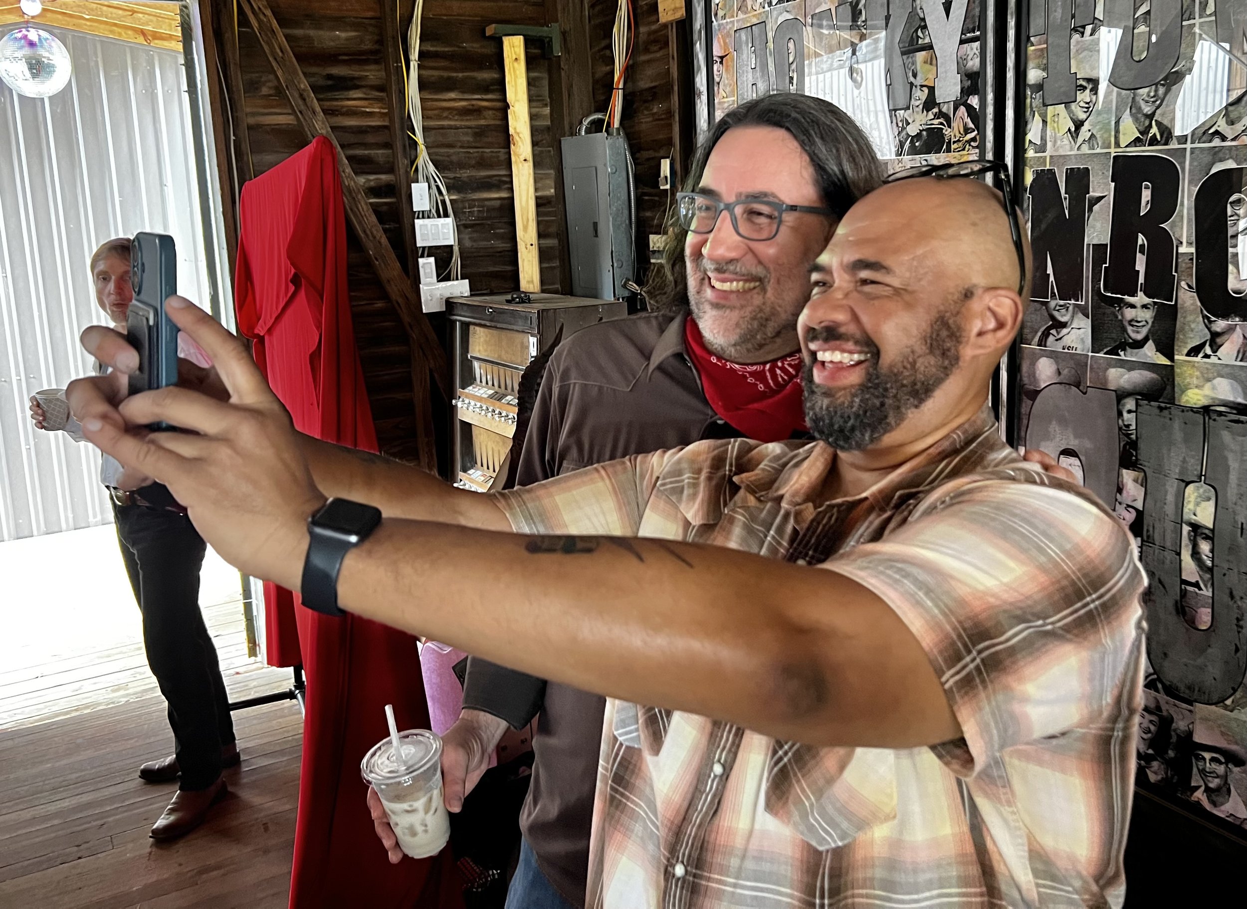 Two men taking a selfie in a rustic indoor space with wooden walls. One man is holding a drink, and a woman in the background holds a cup.