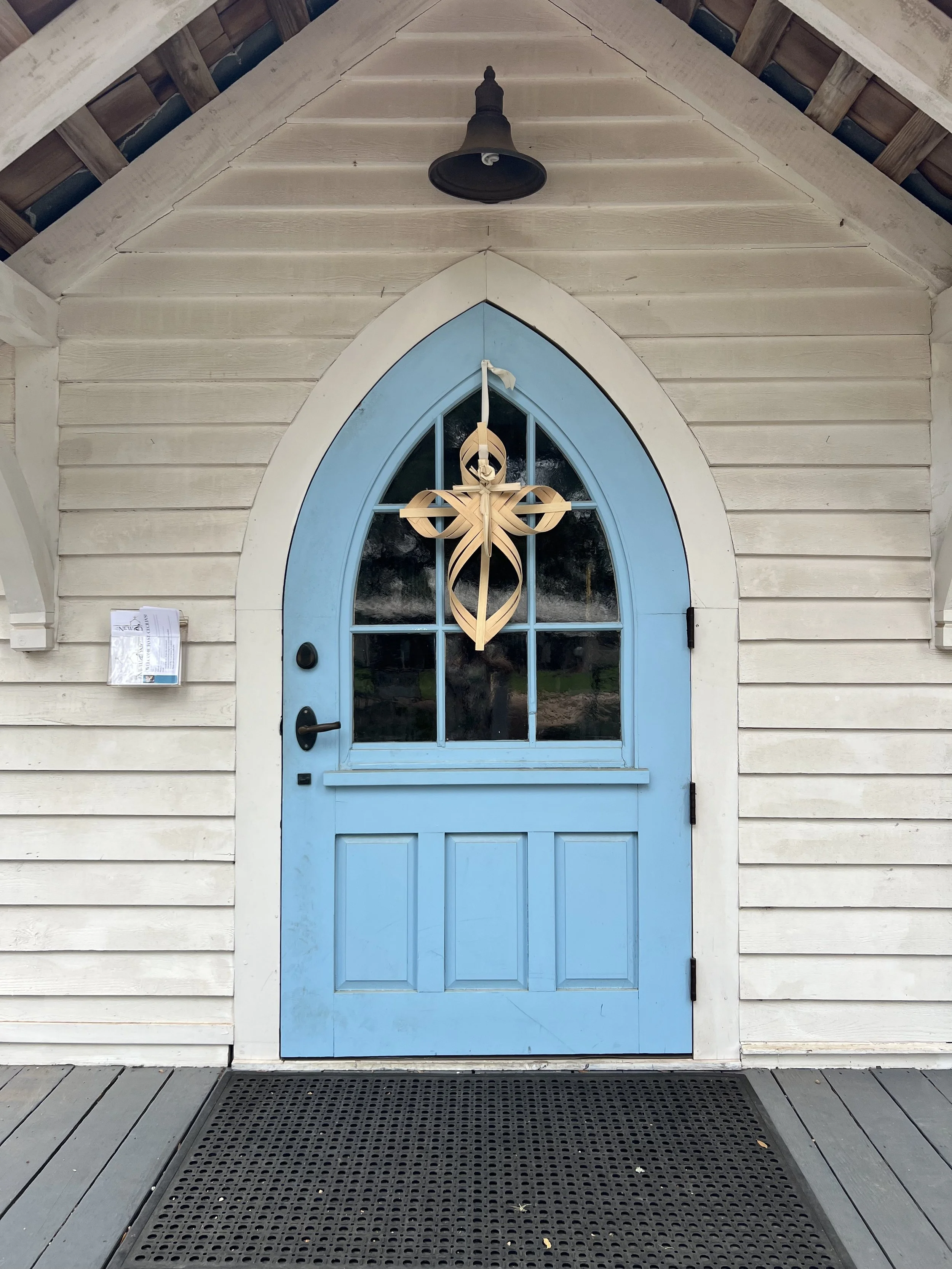A white wooden building with a blue arched door, a black handle, and a decorative cross hanging in the window. There is a black doormat in front of the door, and a roof with exposed wooden beams. A bell or lantern hangs from the roof above the door.