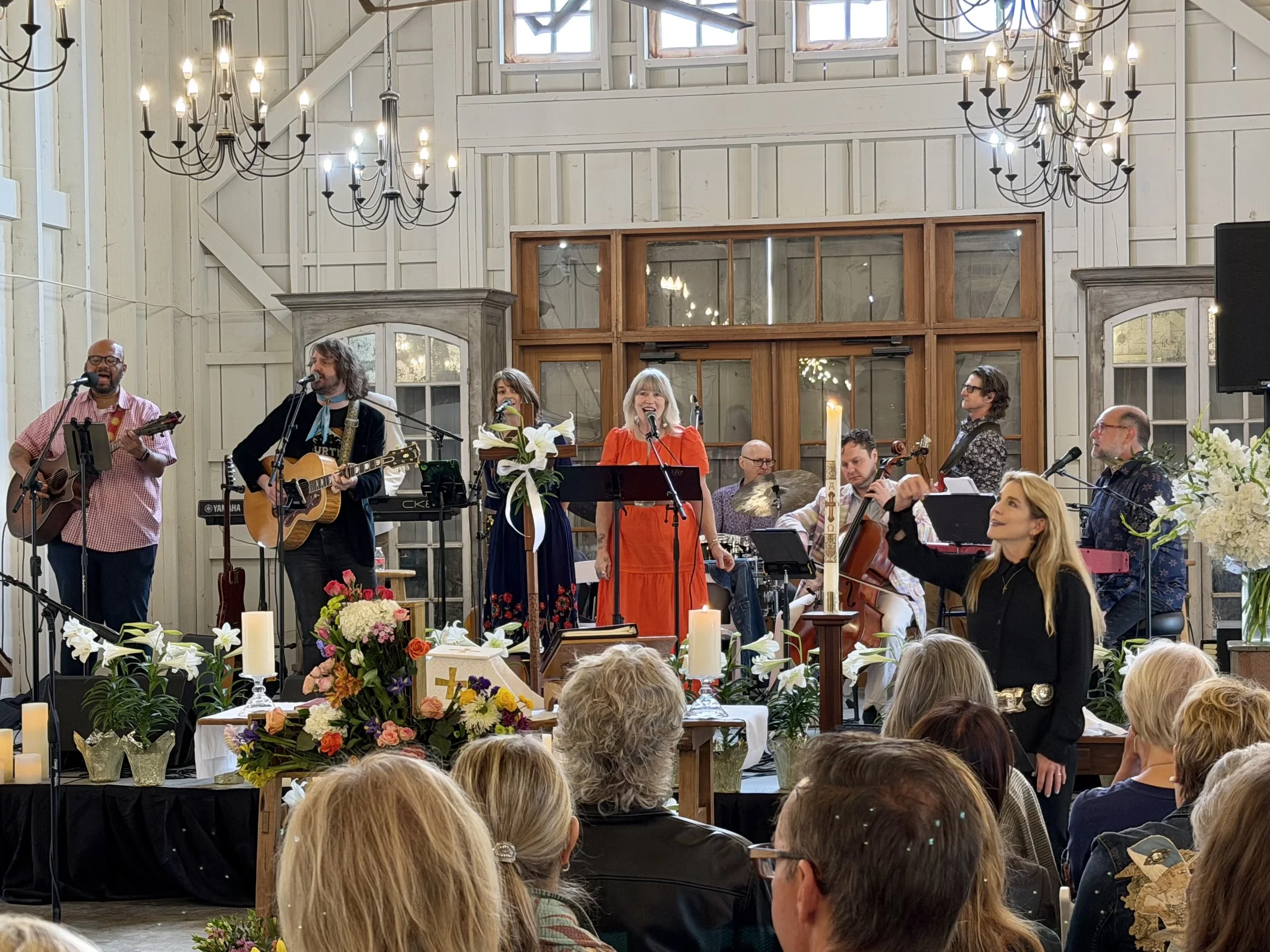 A group of musicians performing at a church service or event with floral arrangements and candles on tables in the foreground.