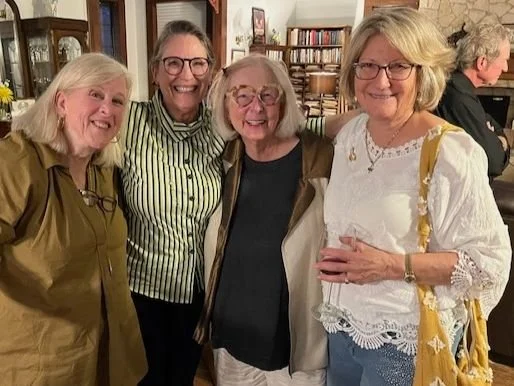 Four women smiling and posing together in a cozy indoor setting, with bookshelves visible in the background.