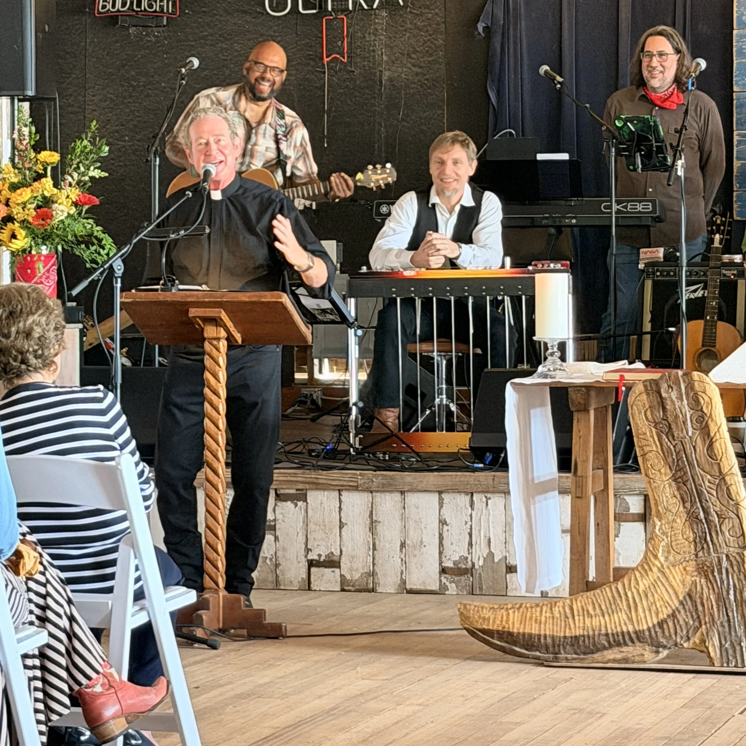 Four men are on a stage with musical instruments, microphones, and a flower arrangement, smiling and performing. One man is singing into a microphone, and another is playing guitar. Two others are seated at a table, smiling. The stage has a sign that