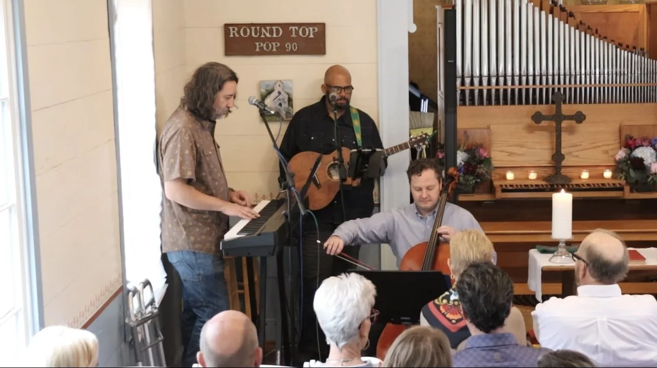 A band performing in a church, with a keyboardist, guitarist, and cellist, in front of an audience. The church features a wooden altar and a large organ with candles and flowers.
