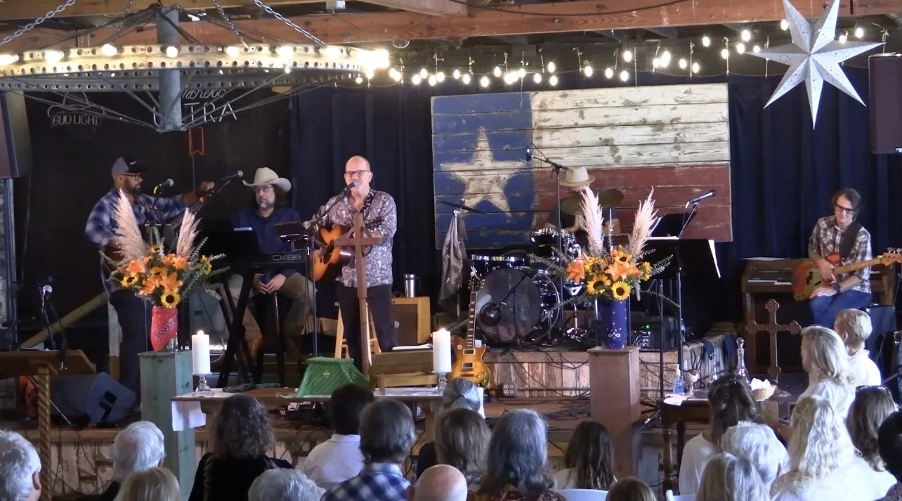 A live music band performing on stage with a rustic, country theme. The band includes five members: a musician with a microphone and guitar, a keyboard player, a singer with an acoustic guitar, a drummer with a hat, and a guitarist. The stage is deco