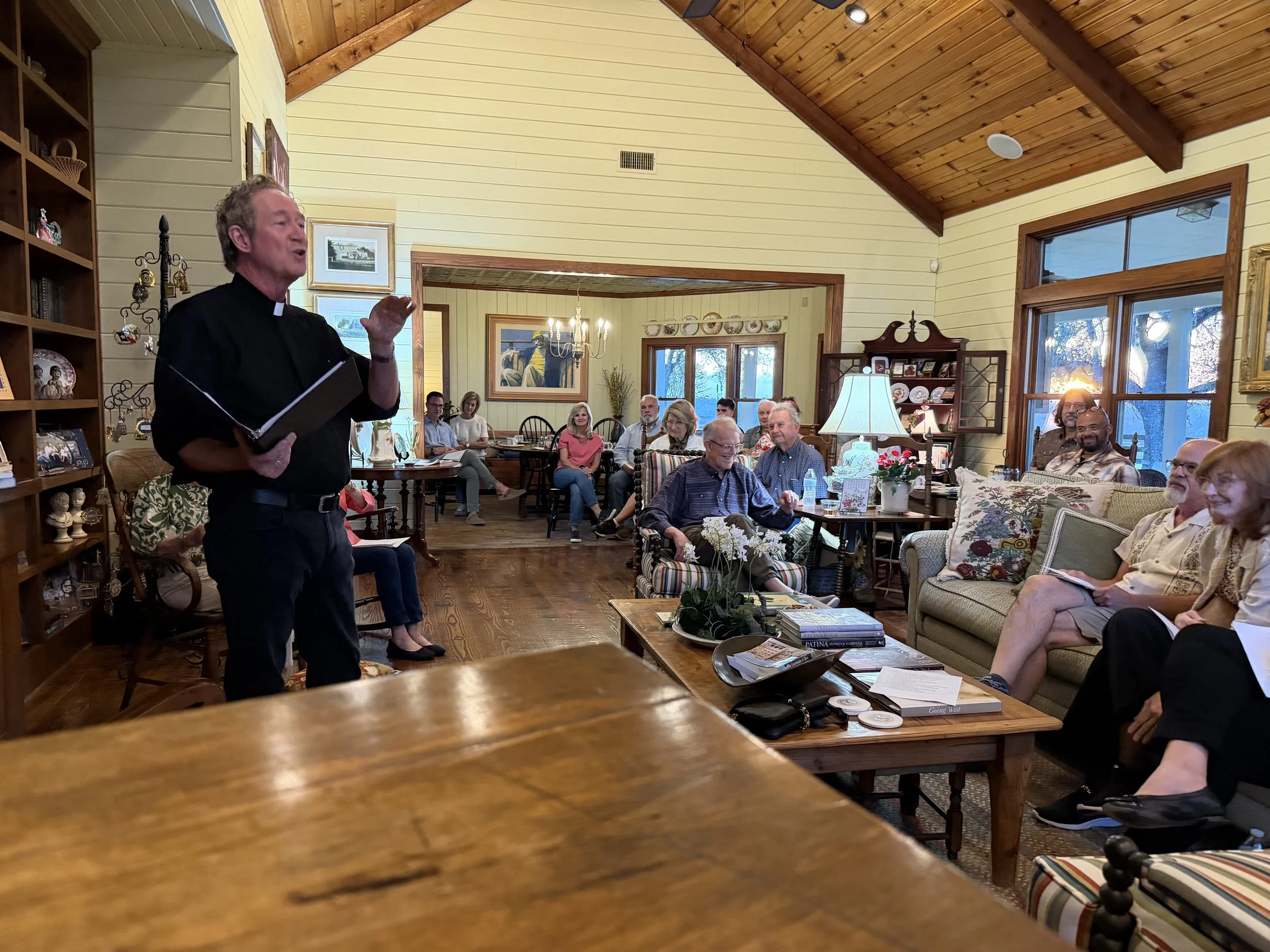 A priest in black clerical attire holding a notebook is speaking to an audience in a cozy living room with yellow walls, wooden ceiling, and large windows. The audience, mainly older adults, are seated on various sofas and chairs, listening attentive