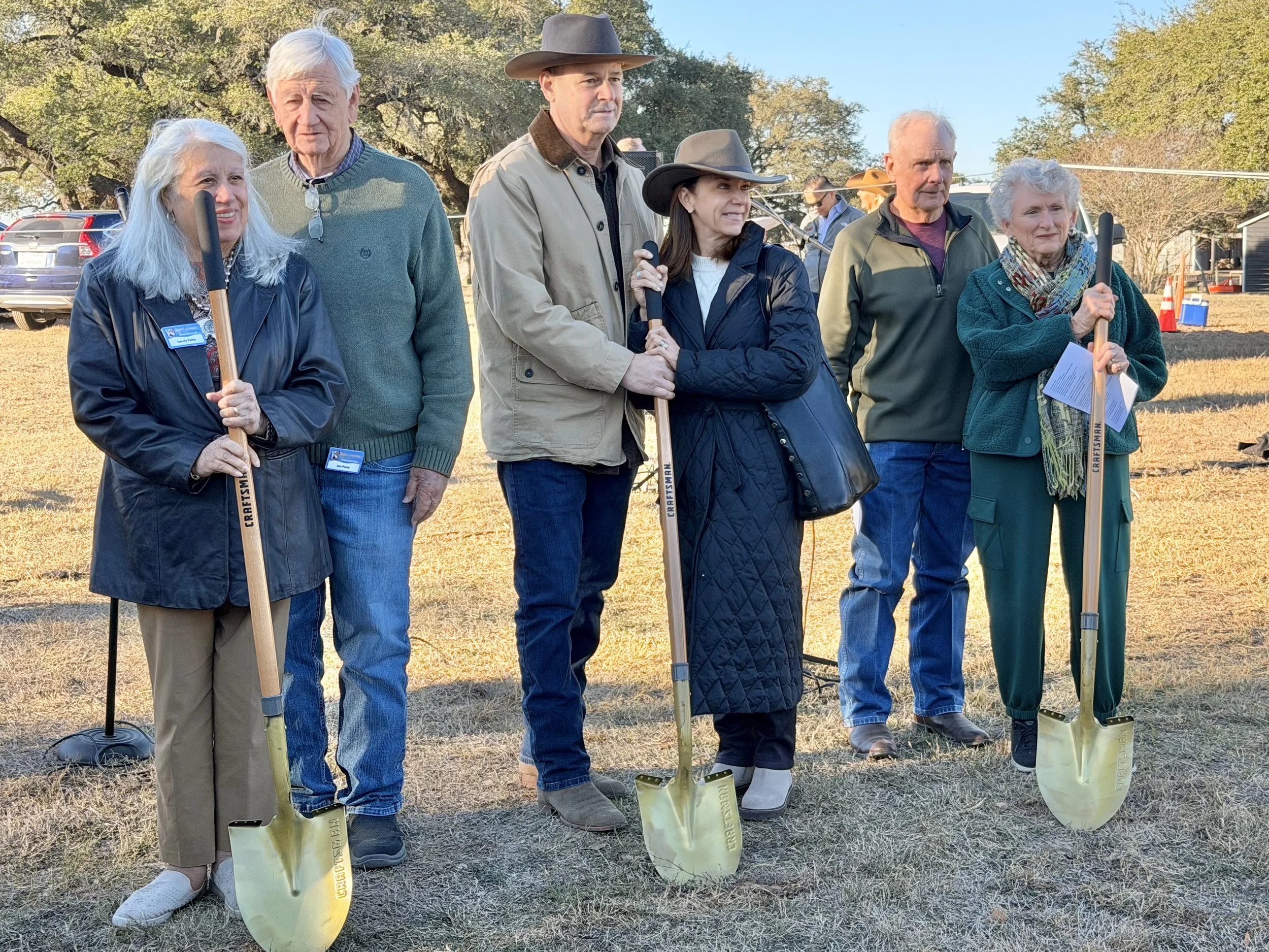 A group of older adults standing outdoors on a sunny day, holding small shovels, with trees and cars in the background.