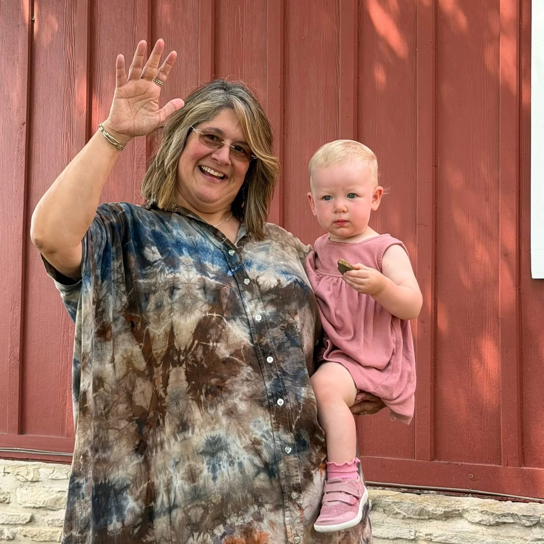 A woman with blonde hair and glasses smiling and waving at the camera while holding a young girl with blonde hair and blue eyes, dressed in pink, standing in front of a red wooden door.