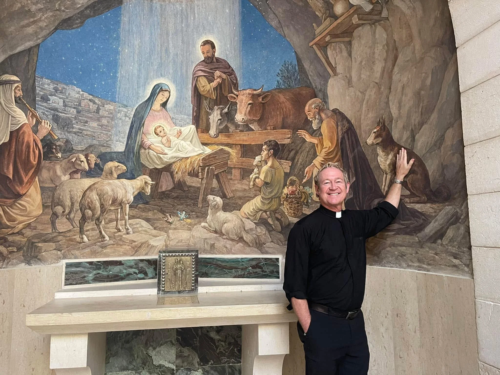 A man dressed as a priest smiling and posing in front of a religious painting depicting the nativity scene, featuring Mary, baby Jesus, Joseph, and farm animals.