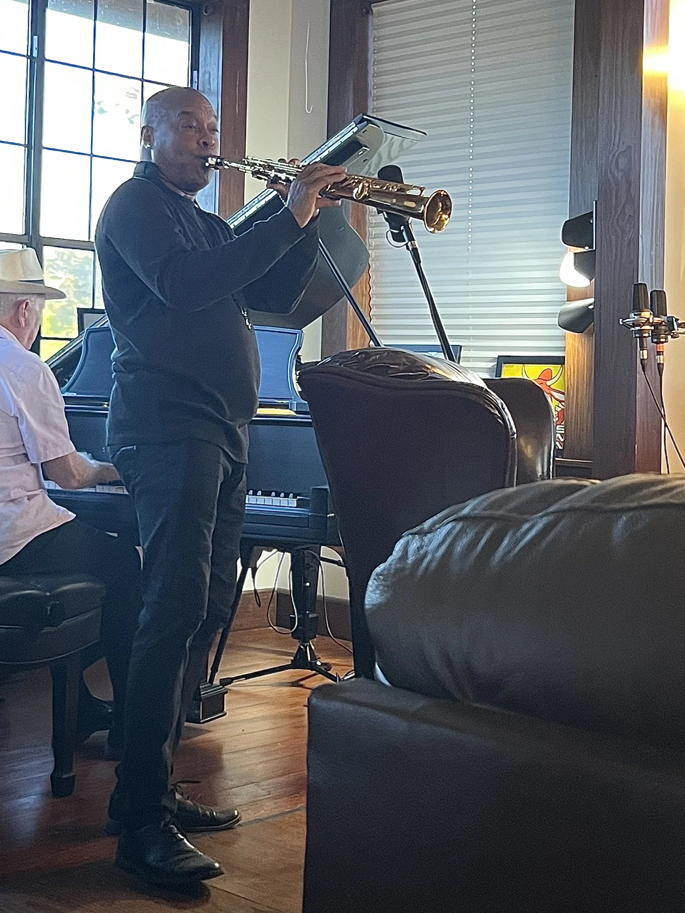 A man playing a saxophone at an indoor event, standing near a microphone with a music sheet in front of him, during daylight with sunlight coming through large windows.