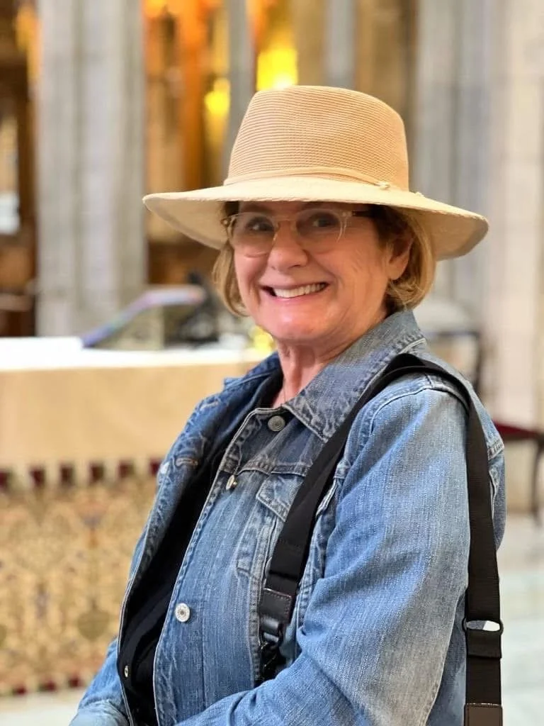 A woman with glasses smiling, wearing a wide-brimmed straw hat, denim jacket, and carrying a backpack, indoors with wooden structures in the background.