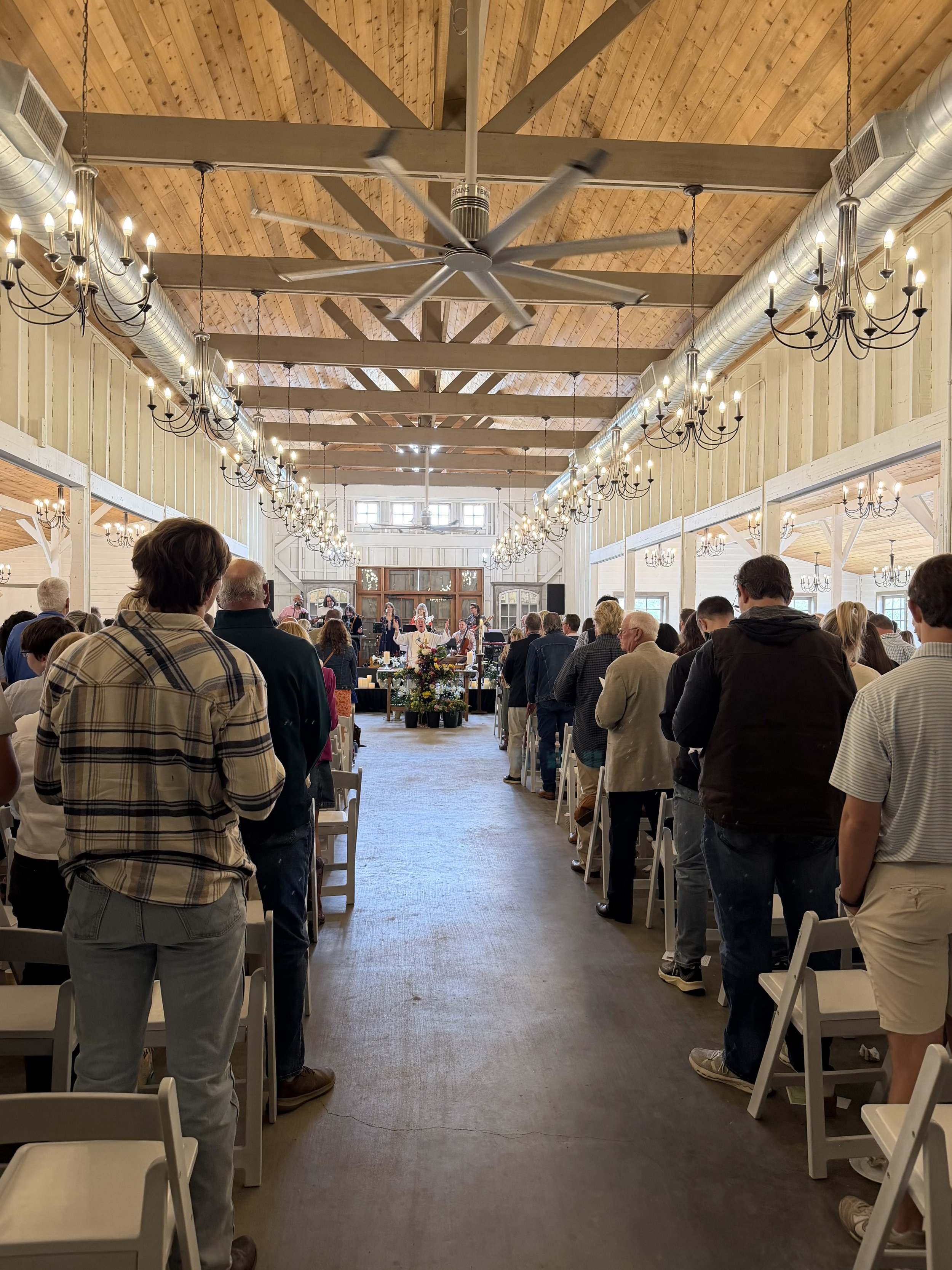 A gathering of people attending a service or event inside a spacious, rustic venue with high wooden ceilings, chandeliers, and a large ceiling fan. The audience is standing and facing a stage at the front, where a band or speaker is present.