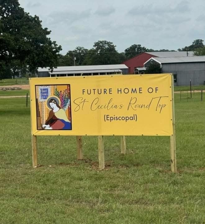 Yellow sign with an illustration of a woman with dark hair, a face mask, and colorful feathers in her hair, seen sitting in a stained glass window. The sign says 'Future Home of St. Cacilia's Round Top (Episcopal)' and is placed on a grassy area with