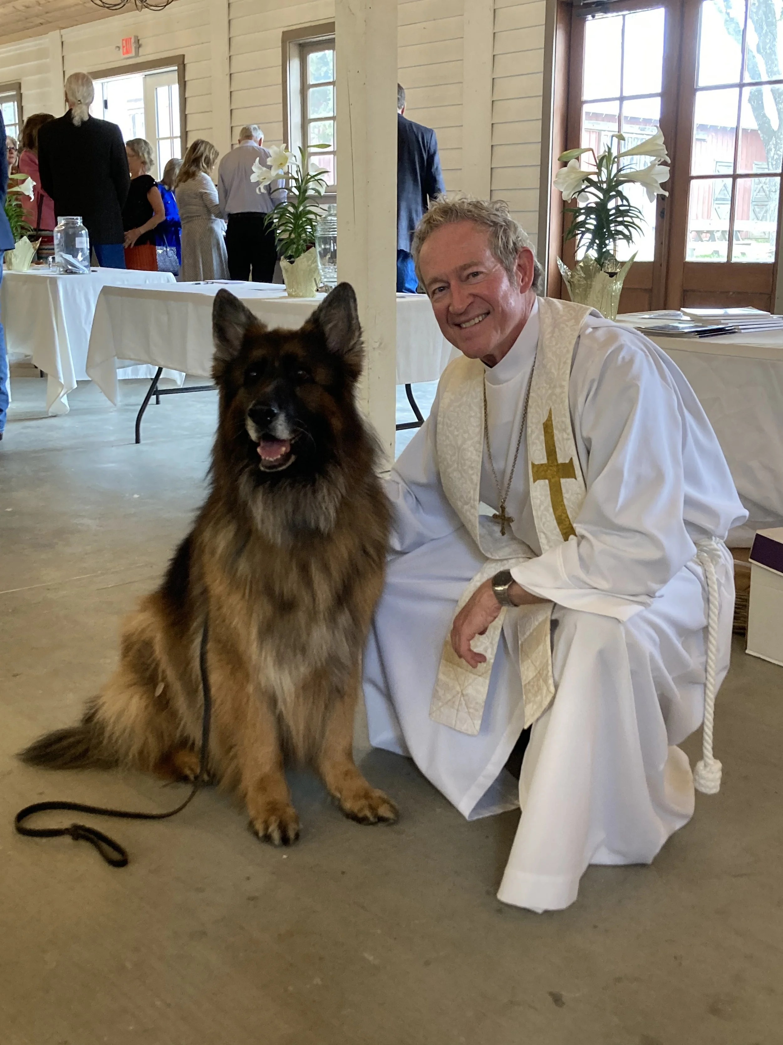 A smiling man dressed in religious attire with a gold cross necklace, kneeling beside a large dog with a thick multicolored coat, inside a wooden hall with several people and floral decorations in the background.