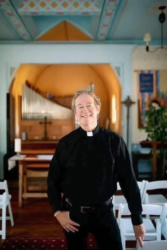 A smiling male priest in black clerical clothing standing inside a church with wooden furnishings and colorful decorations, including a pipe organ and plant.