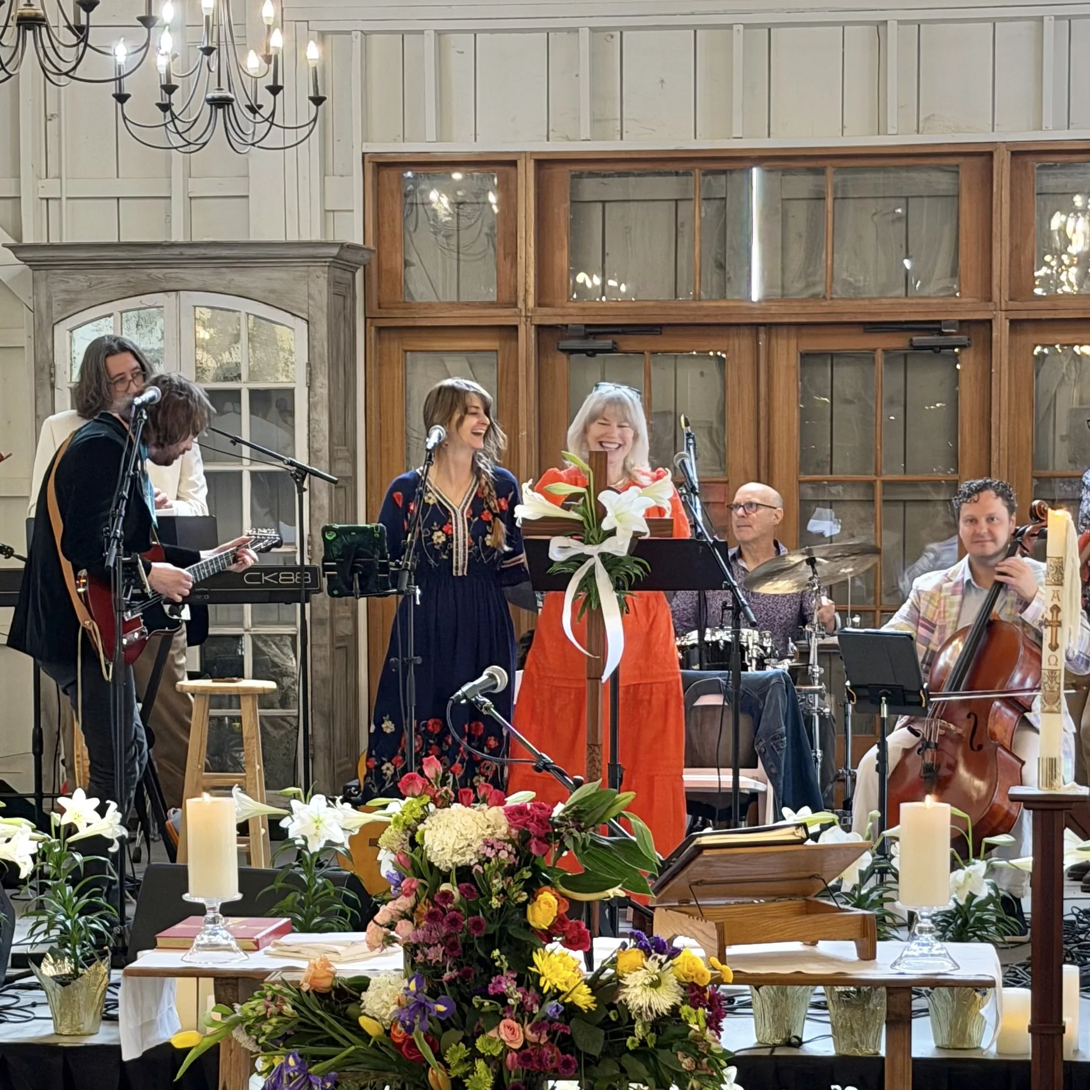 People gathered at a celebration or event indoors with floral arrangements on the table and a view of a large window and a chandelier hanging from the ceiling.