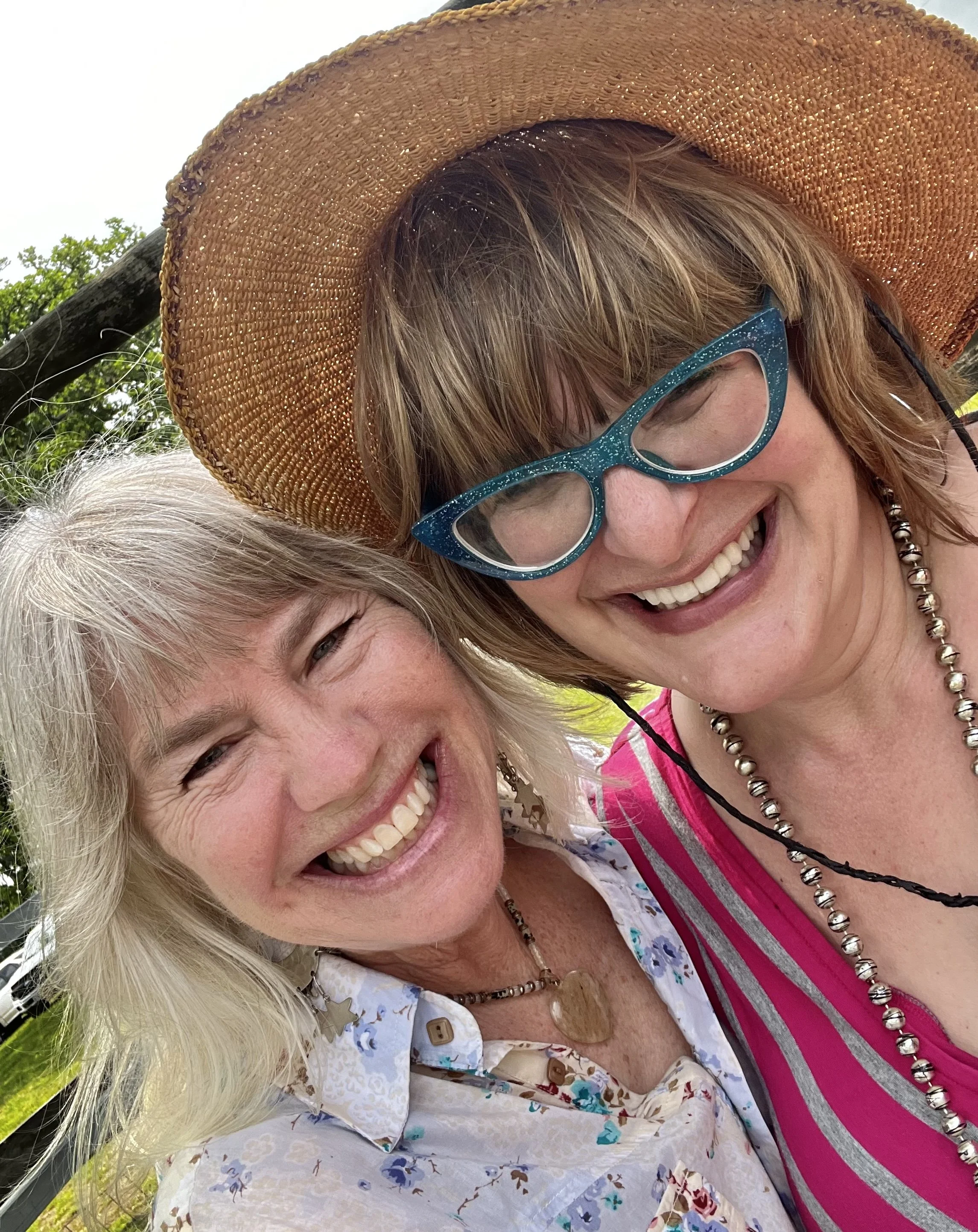 Two women smiling and smiling with their faces close together outdoors. One woman has blonde hair, glasses, a large straw hat, and a floral shirt; the other has light-colored hair and is wearing a striped pink and gray top with a beaded necklace.