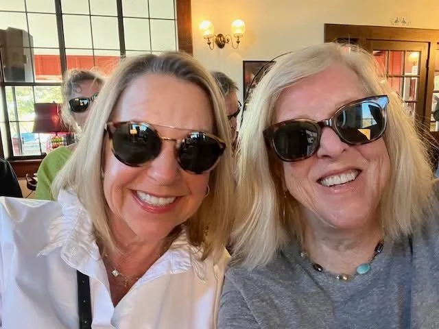 Two smiling women wearing sunglasses taking a selfie inside a cozy cafe or restaurant with warm lighting.