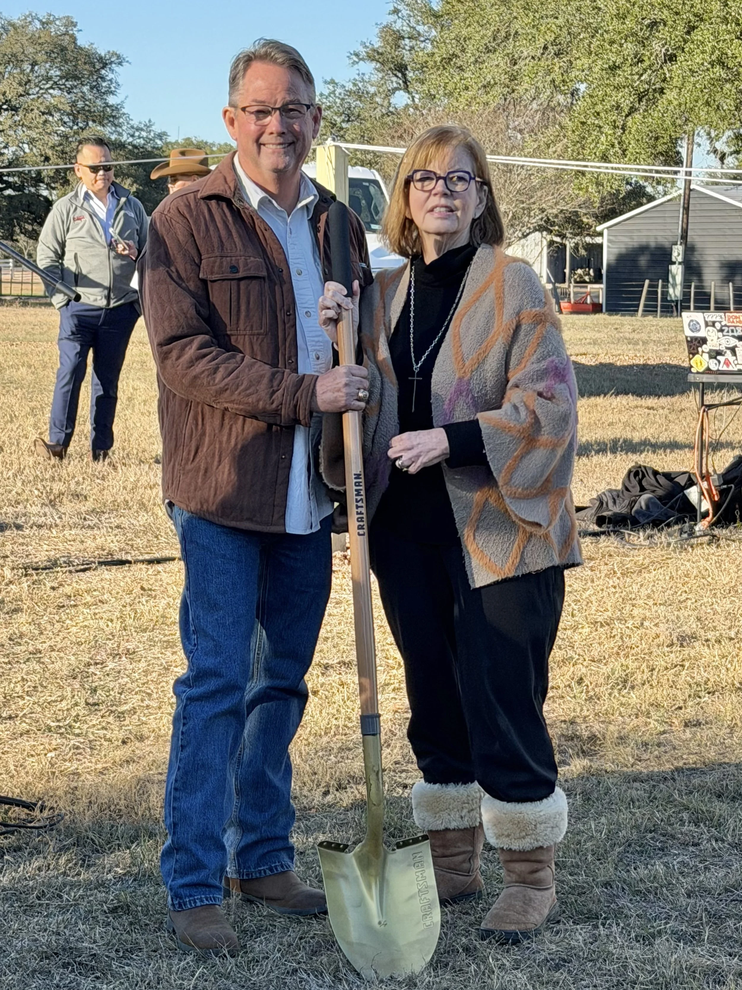 Two people, a man and a woman, standing outdoors on grass with trees in the background. The man is holding a yellow shovel with a wooden handle. The woman is wearing a colorful coat and cozy boots. There are two men in the background, one wearing sun