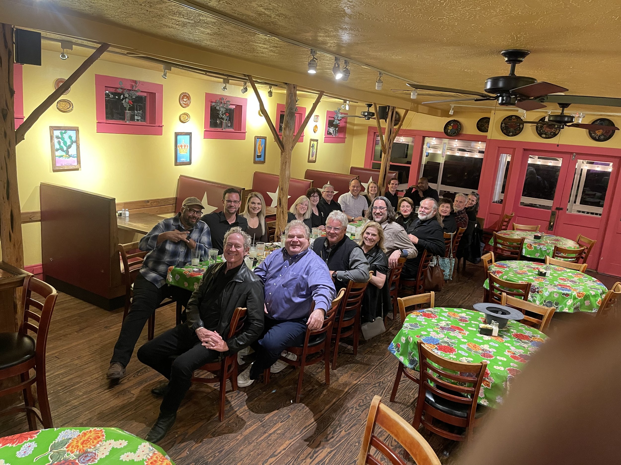 Group of people sitting around a table in a restaurant decorated with colorful artwork, pink window frames, and festive tablecloths.