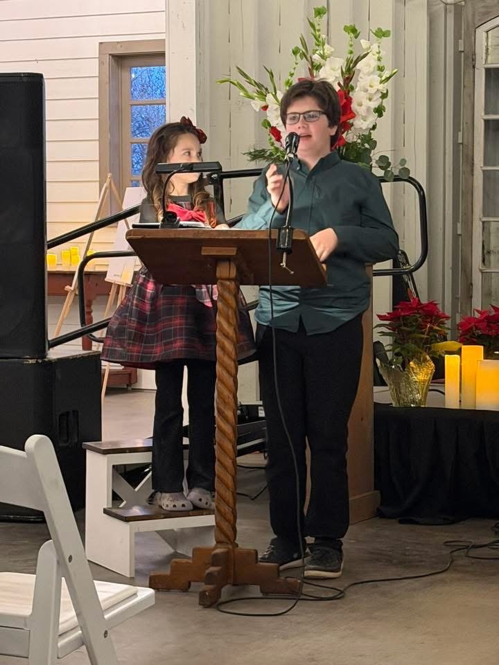 A young boy and a girl are standing at a wooden podium with a microphone, speaking or singing. The girl is on a small step stool, wearing a plaid skirt and gray Crocs, while the boy is wearing glasses and a dark shirt. Behind them are large white and