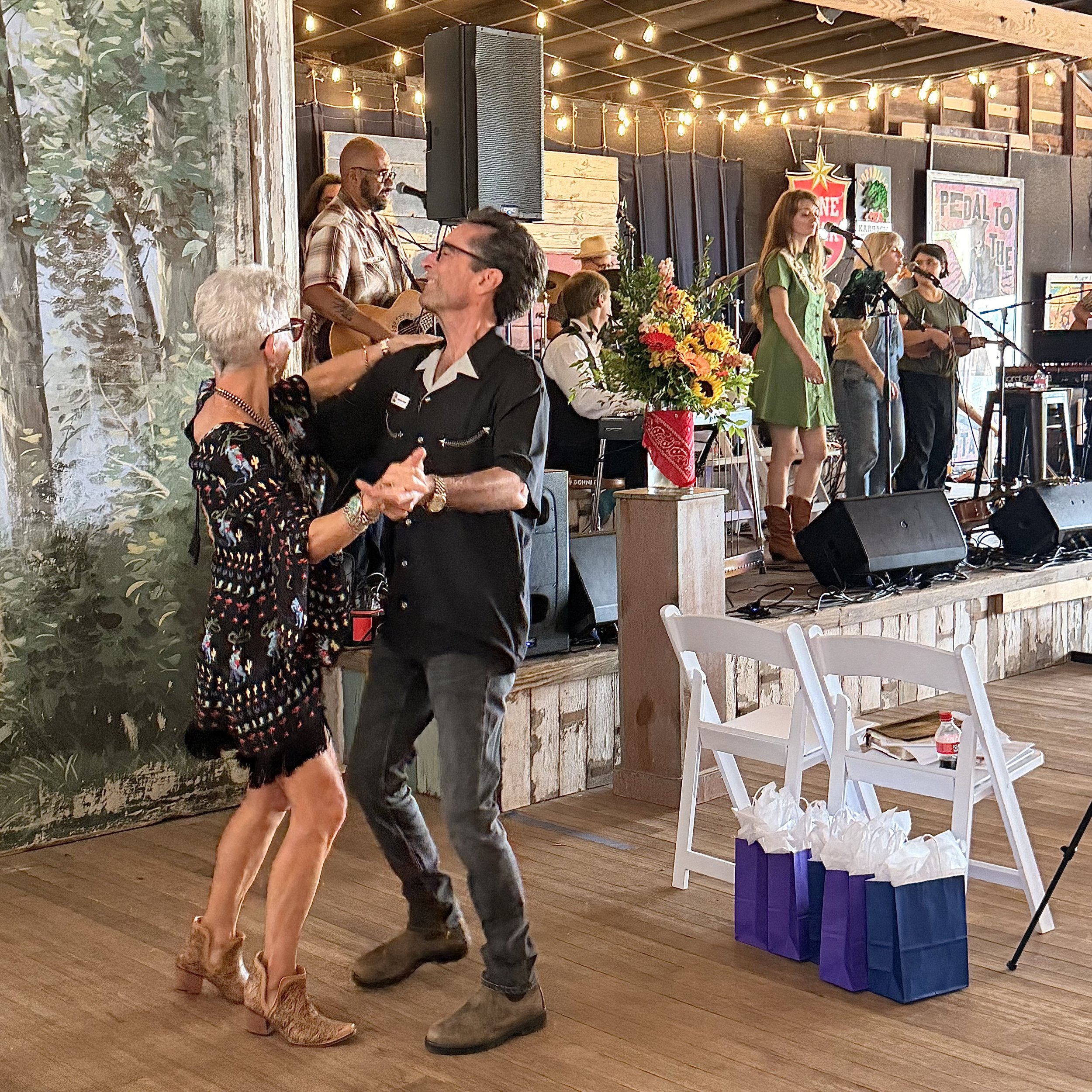 A man and woman are dancing together, holding hands, in a lively indoor setting. There is a band performing on a stage in the background, with members playing guitars and singing. The room is decorated with string lights, flowers, and posters on the 