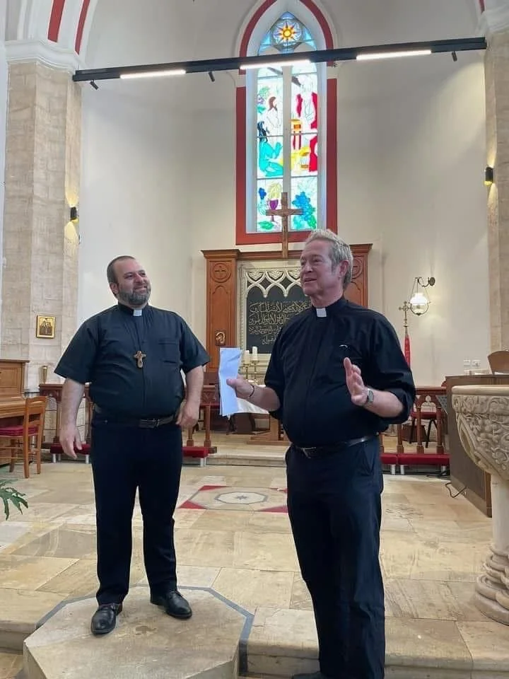 Two male clergy members in black clergy shirts standing inside a church, with one holding a piece of paper and speaking, while the other listens, in front of a wooden altar and stained glass windows.