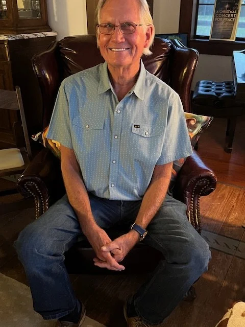 An elderly man with blonde hair and glasses sitting in a leather armchair, smiling, indoors in a cozy room with wooden floors and a window in the background.