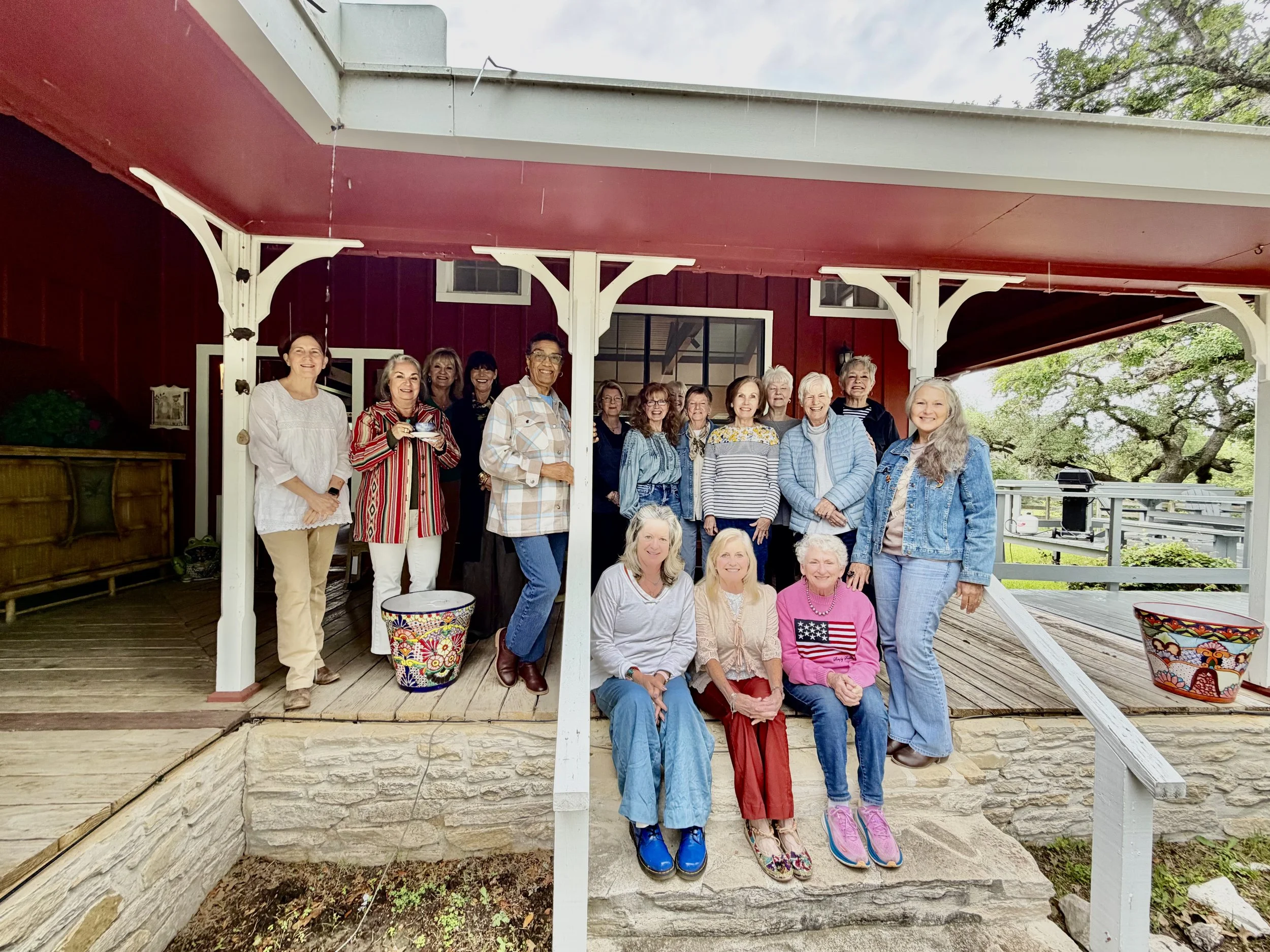Group of women gathered on a porch with trees in the background, some women are standing and some are sitting, smiling for a photo.
