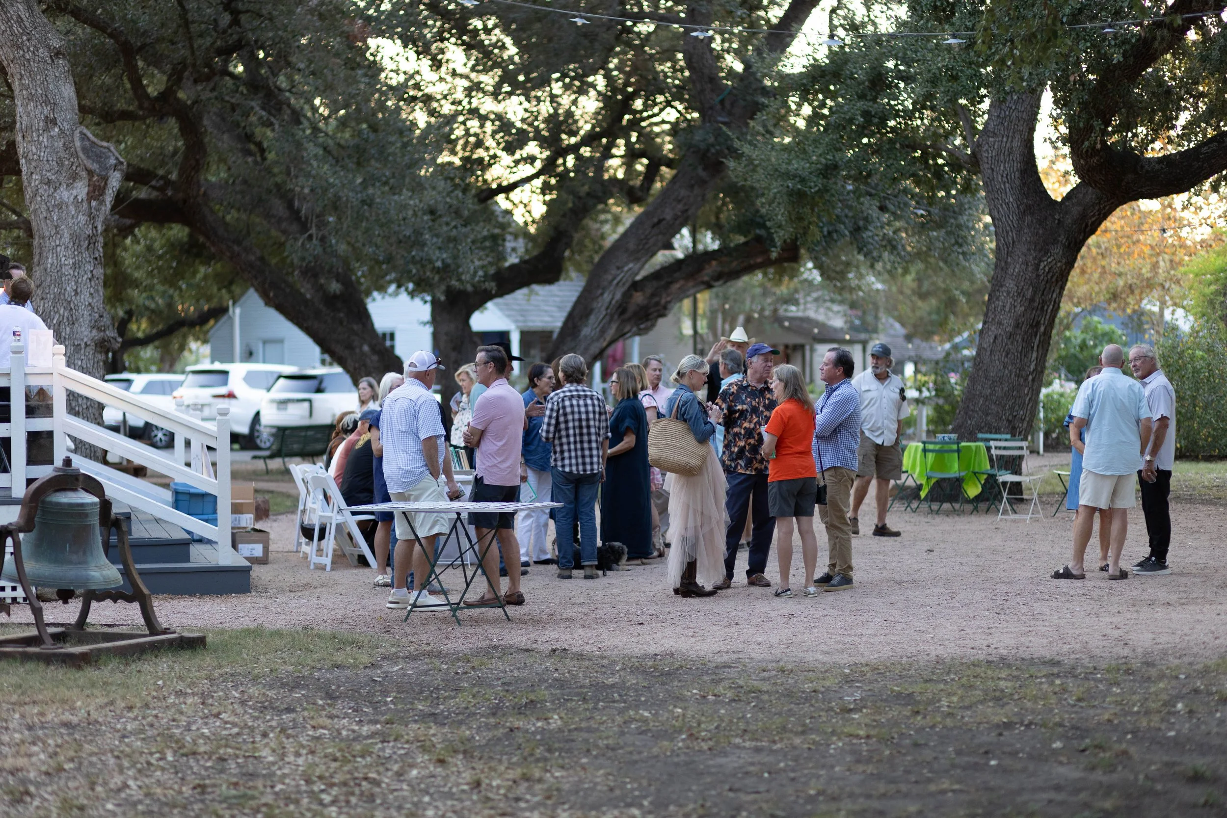 Group of people gathered outdoors in a park, socializing under large trees with houses and parked cars in the background.