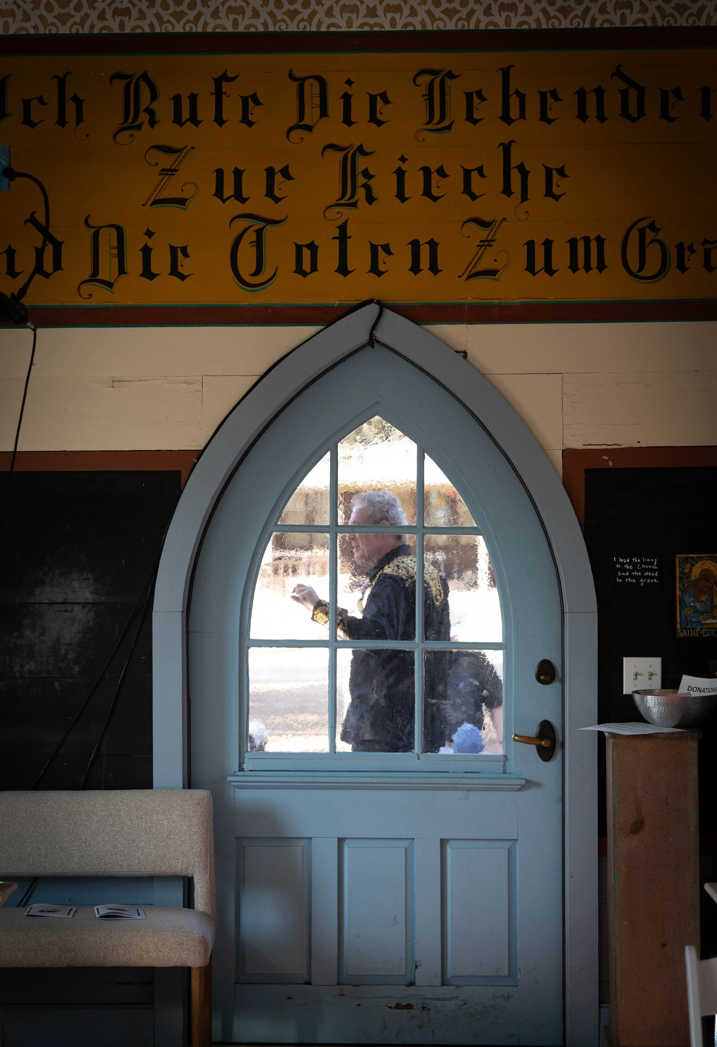 A man in a black jacket with gold embroidery is seen through a decorative glass-paned door inside a church, with a sign in German and an interior with blackboards and a donation bowl.