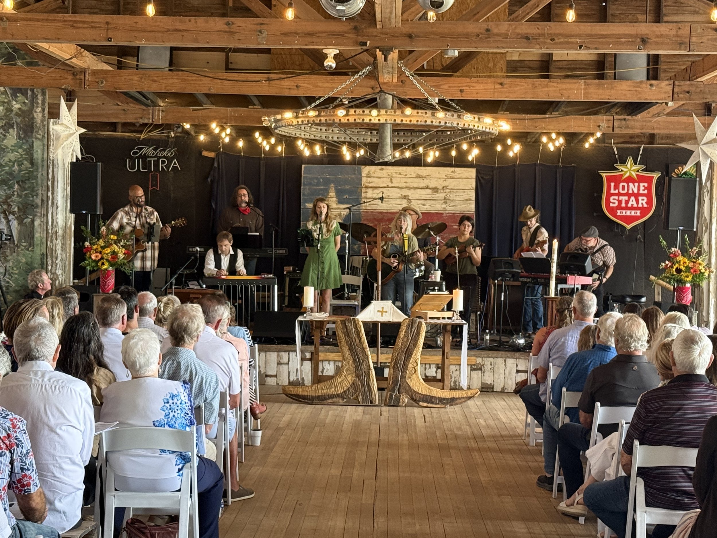 A band performing on stage at an indoor event with audience seated in front. The stage has a rustic wooden backdrop and string lights hanging above.