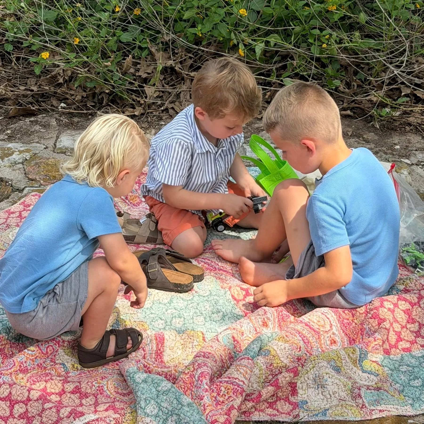 Three children sitting on a colorful quilt outside, examining something with a green bucket nearby, against a background of green plants and dry leaves.