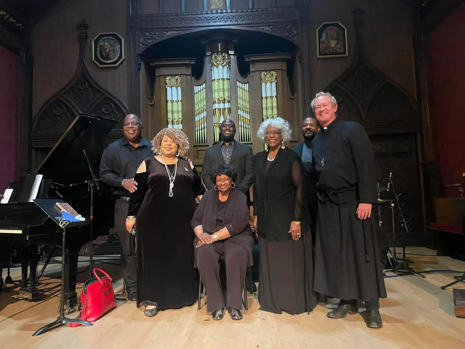 Group of seven people, six standing and one sitting, dressed in black attire, posing for a photo in a concert hall or church with stained glass windows and organ pipes in the background.