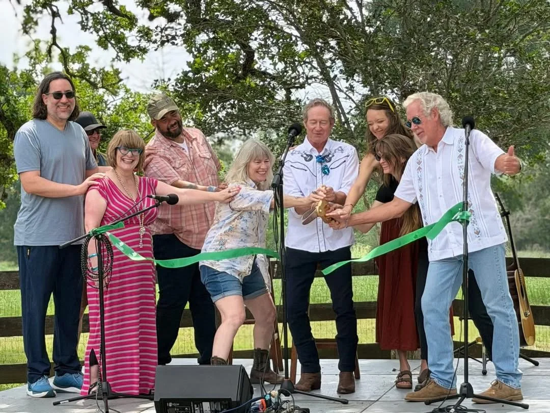 A group of people gathered on a stage outdoors, participating in a ribbon-cutting ceremony. They are smiling and holding a pair of scissors together. The scene includes microphones, a guitar, and trees in the background, indicating a celebration or e