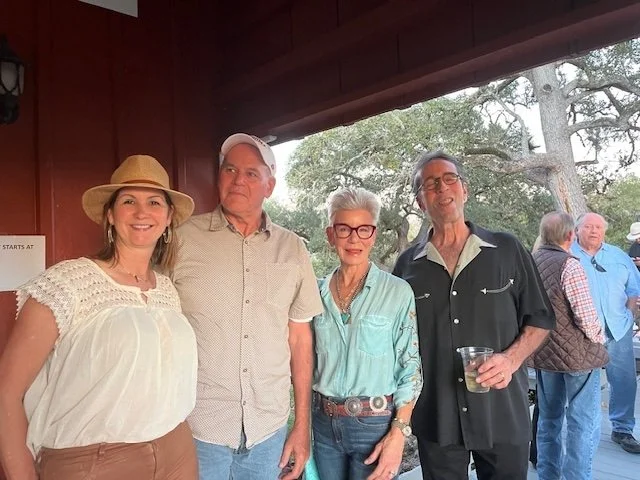 Four people standing together at an outdoor event, smiling, with a background of trees and other attendees.