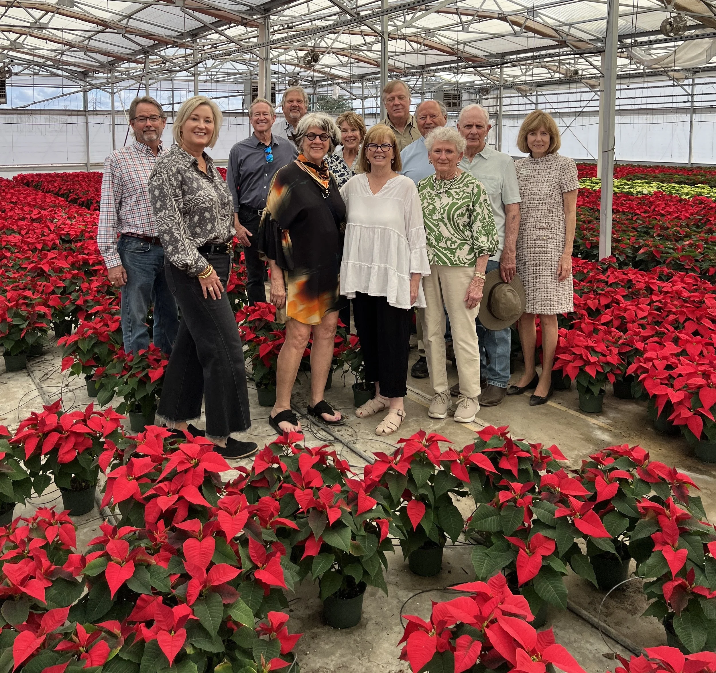 A group of mostly older adults posing together inside a greenhouse filled with red poinsettia plants. They are smiling and standing among the plants, with a metal and glass structure overhead.
