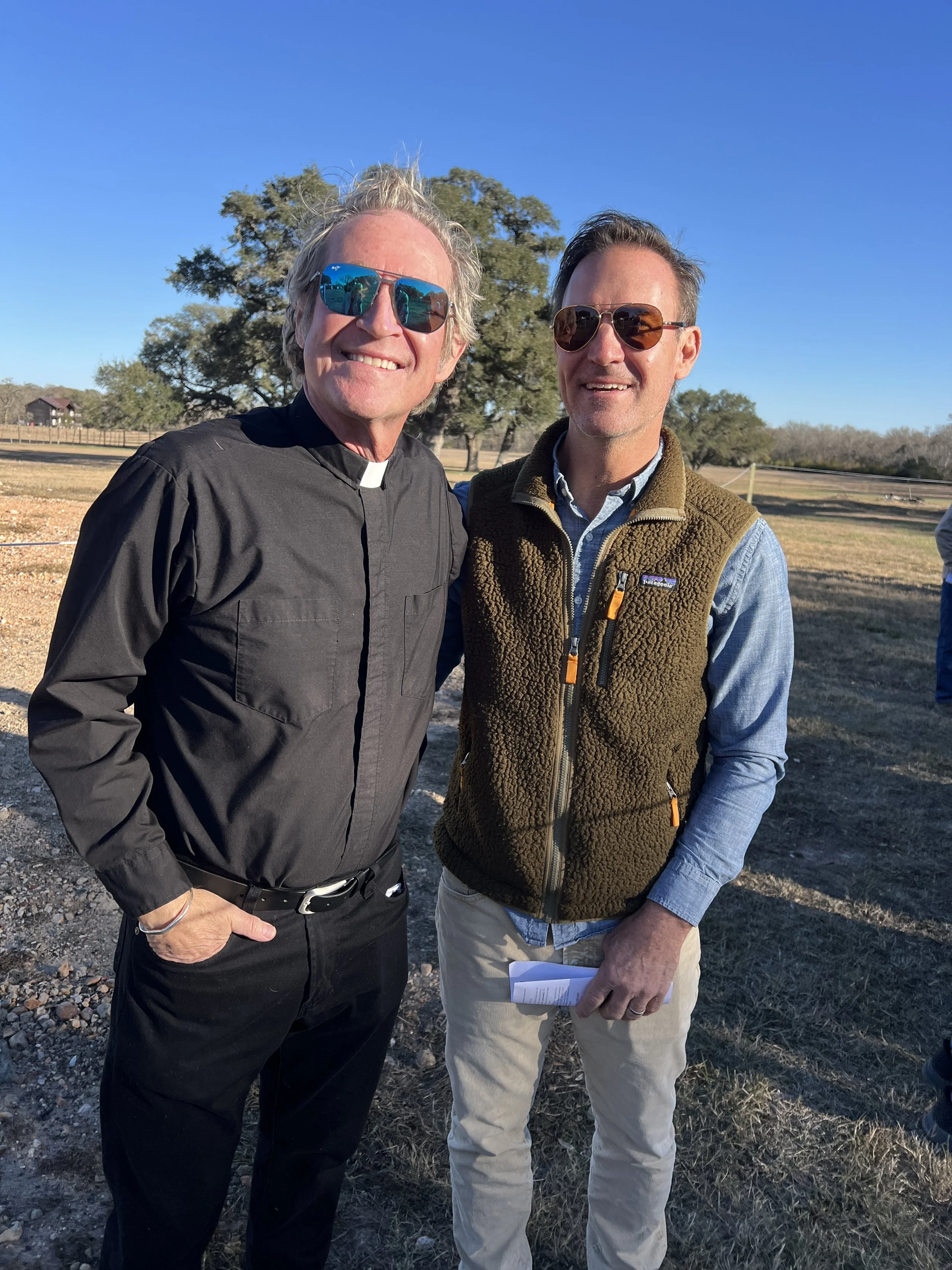 Two men standing outdoors, smiling for a photo. One is dressed in a black clergy shirt with a white collar and sunglasses, and the other is wearing a blue shirt with a brown fleece vest and sunglasses. The background features a large tree, open field