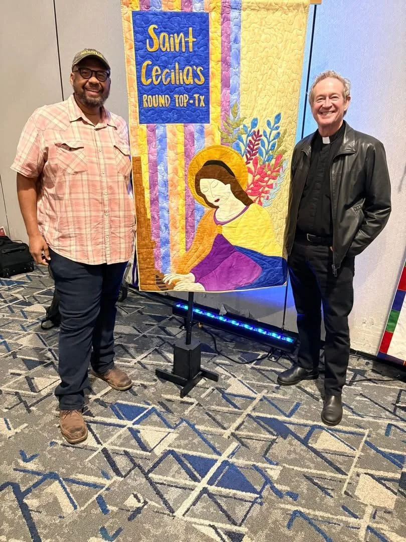 Two men standing beside a colorful church quilt with a depiction of Jesus praying and a sign reading 'Saint Cecilia's Round Top TX'
