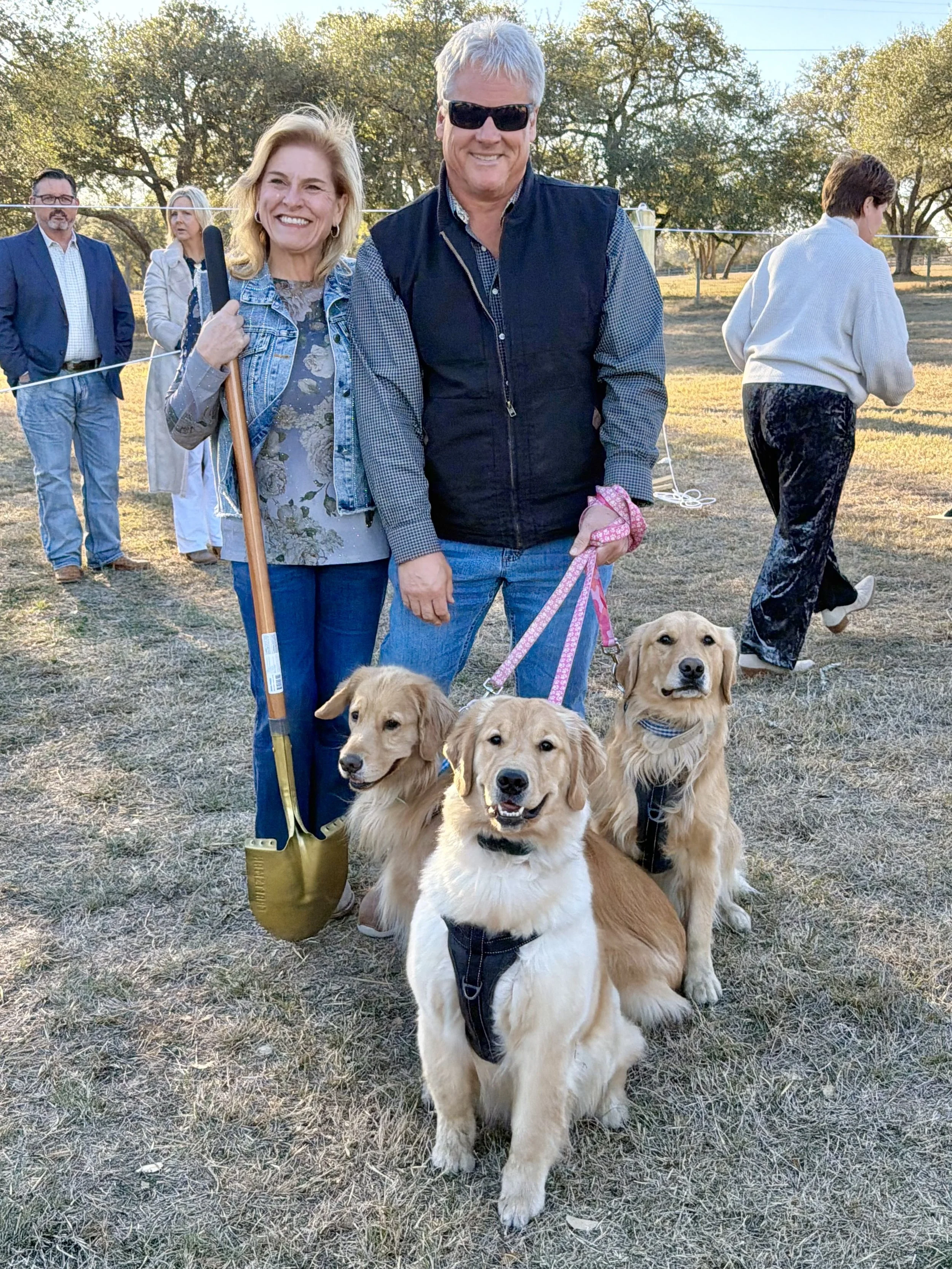 A group of people at an outdoor event with a woman holding a microphone and three golden retrievers in the foreground, set in a grassy area with trees and sunlight.